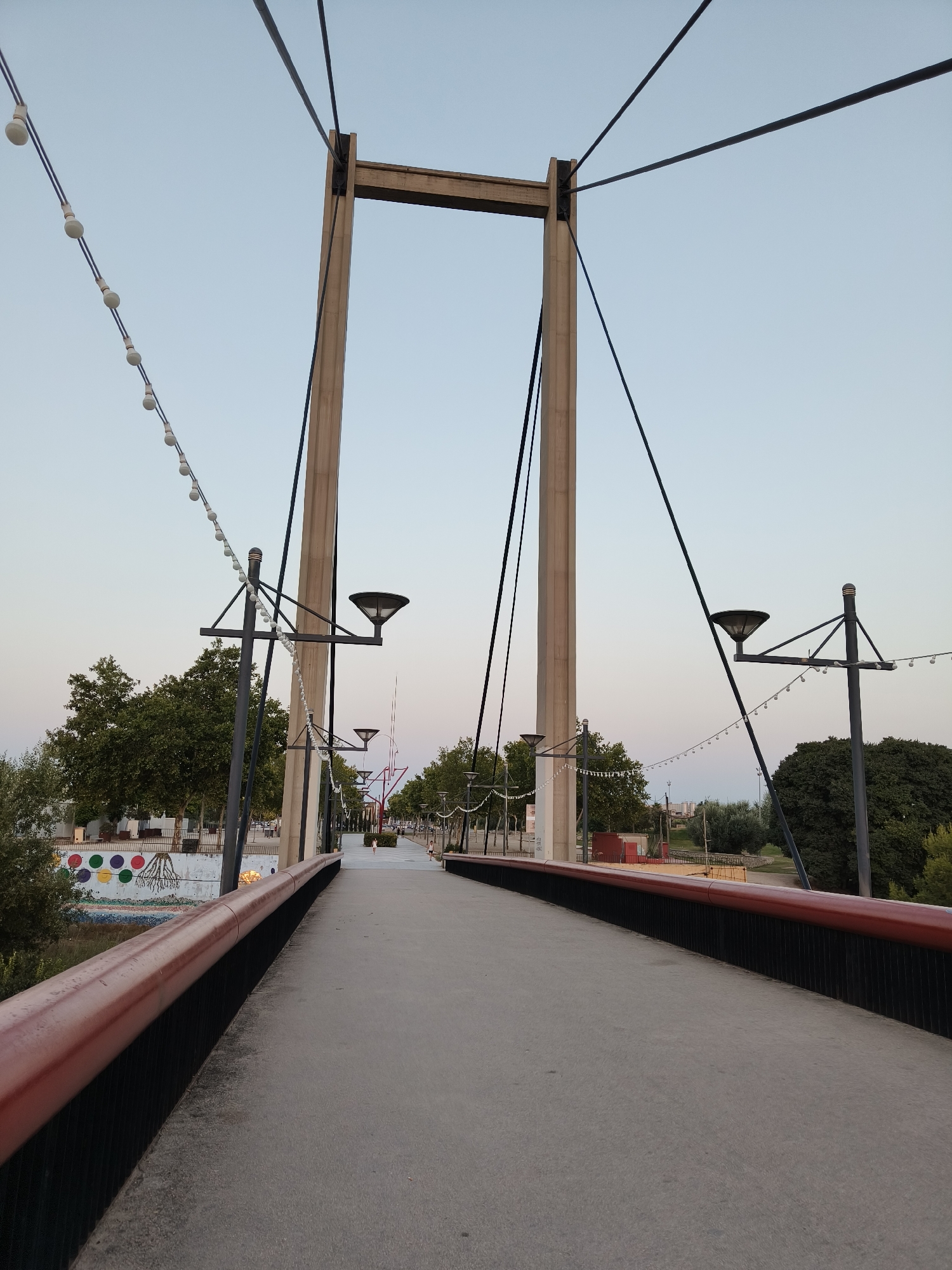 This is a photograph of the Pasarela del Real de Gandia bridge, located in Gandia, Spain. The image depicts a pedestrian walkway with a wide, curved path made of concrete. The walkway is bordered by a dark railing with a reddish-brown top. On either side of the path, there are lampposts with modern, downward-facing lights, and strings of decorative lights are suspended between them.
In the background, large concrete pylons rise vertically, supporting thick cables that stretch across the frame. These pylons are part of the bridge's suspension system. To the left of the bridge, there are trees, a wall with colorful circular murals, and some buildings. To the right, more trees are visible. The sky above is clear and a pale blue, suggesting it is either early morning or late evening. There are no people clearly visible on the bridge itself, though a few figures appear in the distance further along the walkway. The overall impression is one of a tranquil urban environment designed for pedestrian use.