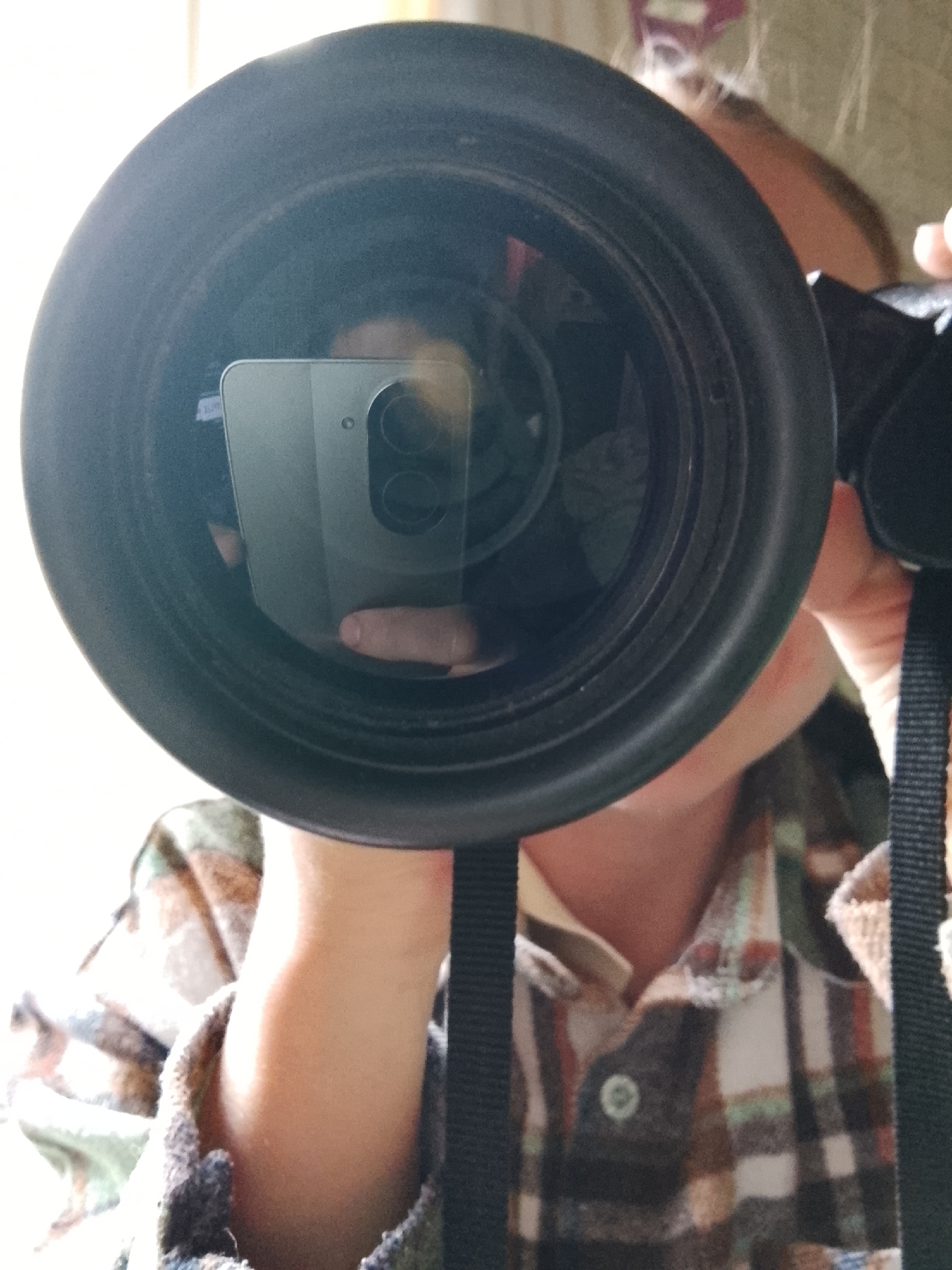A person wearing a plaid shirt is holding a black camera with a large lens in front of their face, capturing a selfie. The reflection in the lens shows the person holding a smartphone, likely the device being used to take the picture. The background is out of focus, but the light suggests an indoor setting. There's no information in the image to indicate the location is Высокиничи, Russia.