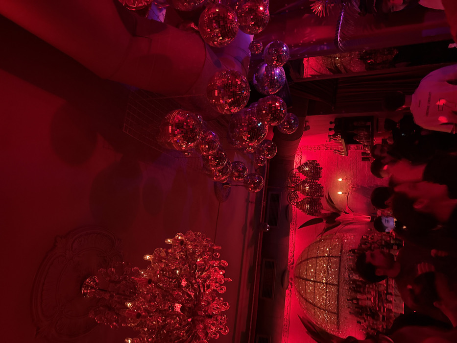 A dimly lit, red-toned interior space features a profusion of reflective disco balls suspended from the ceiling. A decorative chandelier graces the left corner, its intricate design adding to the ambiance. A group of people is clustered on the right side of the frame, partially obscured by shadows. The overall scene suggests a lively party or club environment, enhanced by the red lighting.