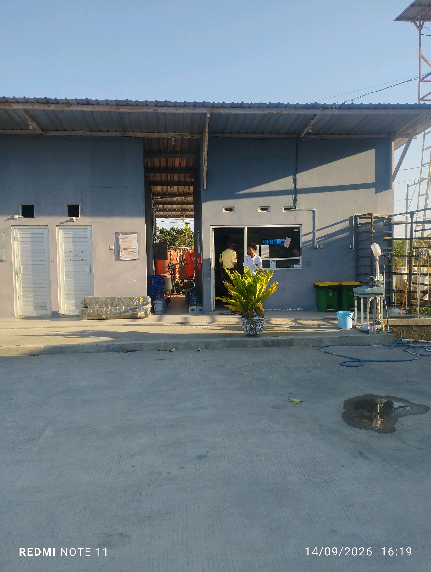 The image depicts a modest, one-story building with a gray exterior and a corrugated metal roof. The building appears to be an industrial or utility facility, possibly a security checkpoint or maintenance shed, given the signage that reads "POS SECURITY" visible on a window. The front of the building features two white doors on the left and an open doorway leading into what looks like a workshop or storage area on the right, where industrial machinery with orange and red components is visible.
In front of the building, a paved area serves as a driveway or parking lot, with a drainage grate and a blue hose lying on the ground. To the right of the open doorway, there are two green trash bins and a blue bucket. A potted plant with large, green leaves sits in front of the security window. Two individuals are partially visible through the security window, one appearing to be wearing a yellow uniform. The scene is illuminated by natural sunlight, suggesting it is daytime. The background reveals trees and a clear blue sky.
The provided information indicates the image was taken in Bero, Indonesia. While the specific location within Bero is not discernible from the visual cues alone, the overall environment suggests a semi-industrial or commercial zone. The timestamp on the image is "14/09/2026 16:19".