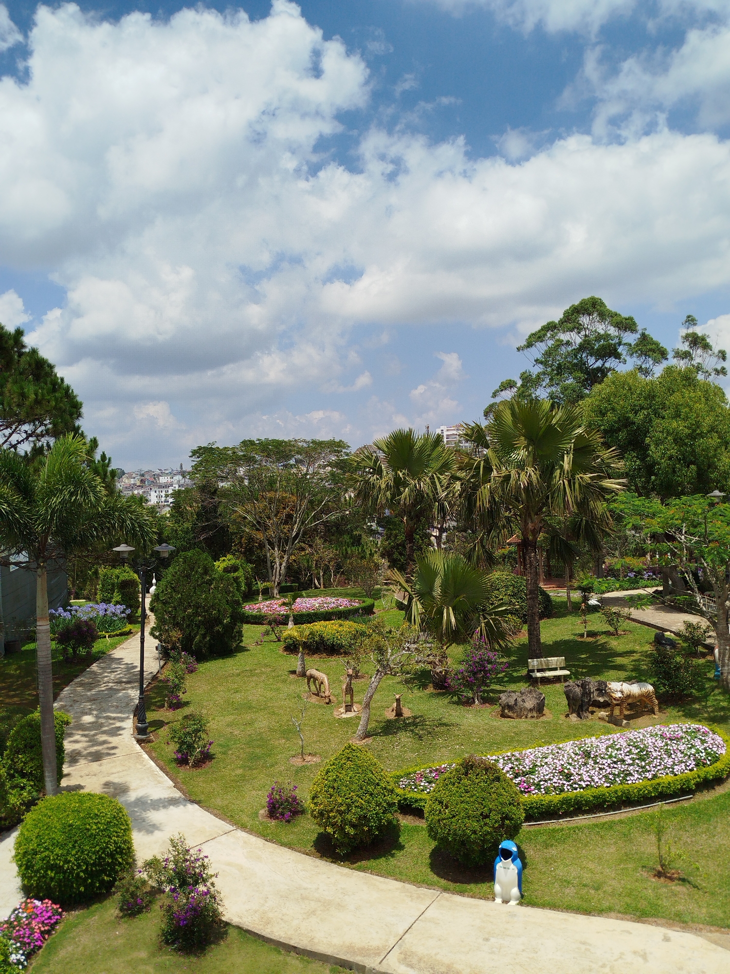 The image depicts a scenic view of a park in Da Lat, Vietnam, characterized by lush greenery and a partly cloudy sky. A winding paved path curves through the landscape, lined with meticulously manicured bushes, trees, and vibrant flowerbeds. In the background, the cityscape of Da Lat is visible, offering a contrast between the natural environment and urban development. The park contains sculptures, benches, and other decorative features, such as a penguin-shaped trash can, enhancing its aesthetic appeal. The overall impression is that of a well-maintained and serene public space, typical of Da Lat's reputation as a city known for its natural beauty and landscaping.