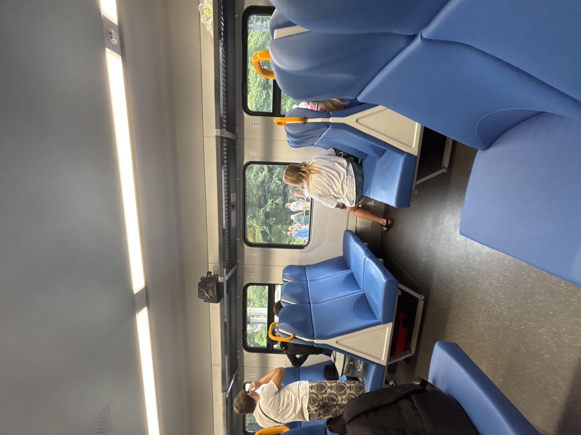 An interior shot of a train car shows rows of blue vinyl seats with white triangular dividers. Overhead luggage racks are visible, with a black bag resting on one. A man in a white t-shirt and patterned shorts stands near a window, holding a phone to his ear. A woman in a white t-shirt and denim shorts sits on a seat facing away from the camera, looking out the window. Outside the window, a group of people can be seen gathered. The floor of the train car is dark gray with a speckled pattern.