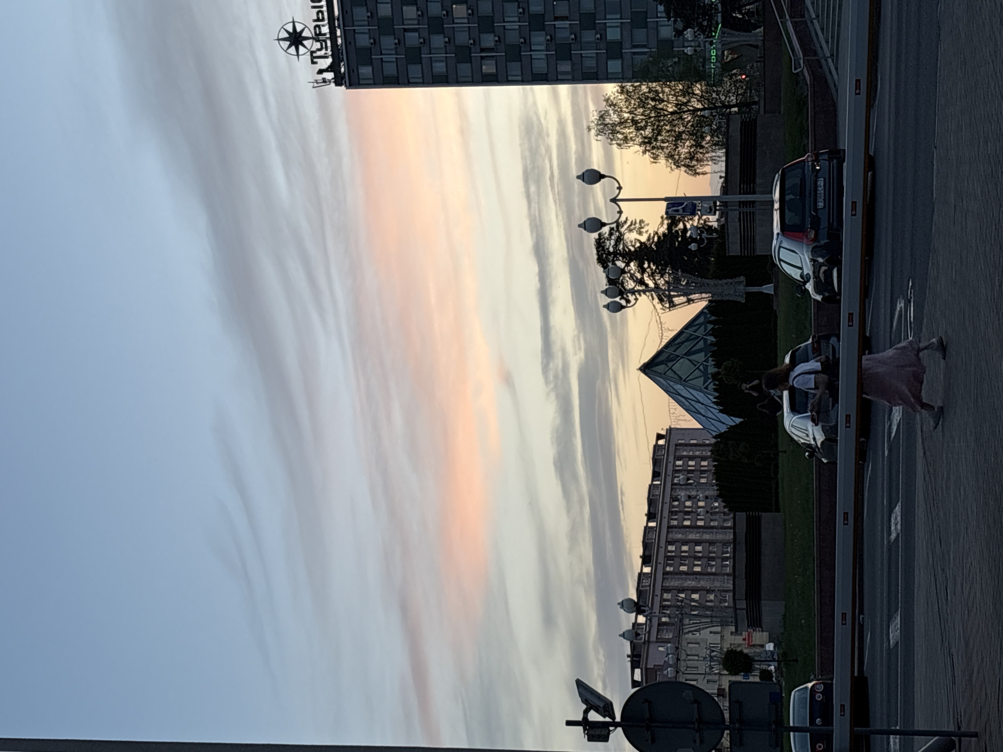 The image depicts a cityscape in Minsk, Belarus, taken at sunset or dusk. The sky is filled with striated clouds, tinged with hues of pink and orange. Tall buildings line the horizon, including a building with a distinctive 'ТУРЫ' sign and a pyramidal glass structure. Streetlights dot the scene. A person in a pink skirt walks along the sidewalk next to a street with cars. A traffic light or a signpost is visible in the foreground.