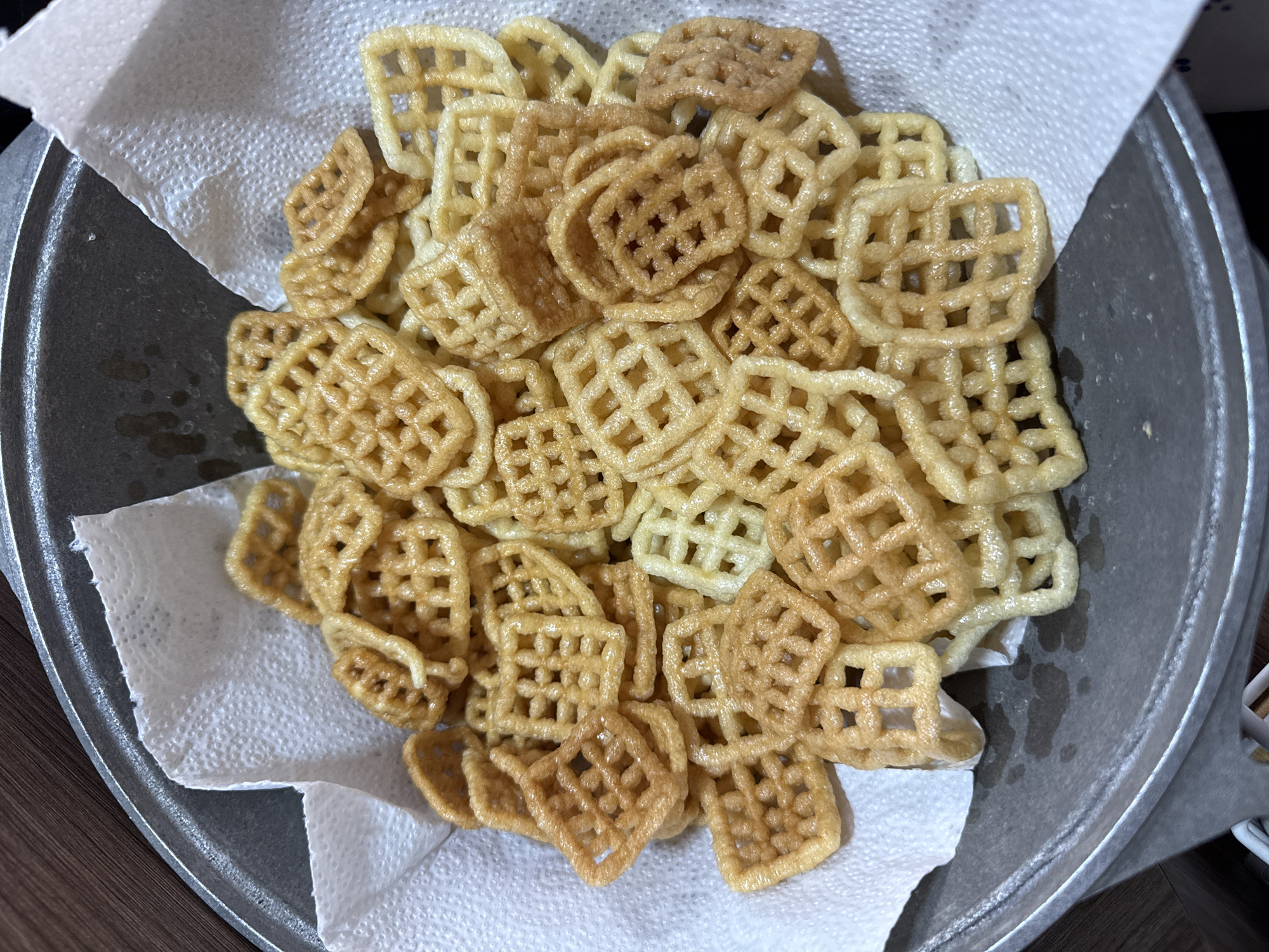 The image shows a close-up, overhead view of a pile of waffle-shaped snacks on a paper towel. The snacks are golden brown and have a lattice-like pattern with square openings. Some snacks appear lighter in color, suggesting they are less fried or a different variety. They are resting on a white paper towel, which is placed inside a shallow, metallic pan. The pan has a slightly worn appearance, and there are some visible oil spots on its surface. The background is blurred, but the texture of a wooden surface is discernible. The lighting suggests an indoor setting, possibly a kitchen, with the light source coming from above. There are no people or text visible in the image. The focus is entirely on the food item, highlighting its texture and appetizing appearance.
