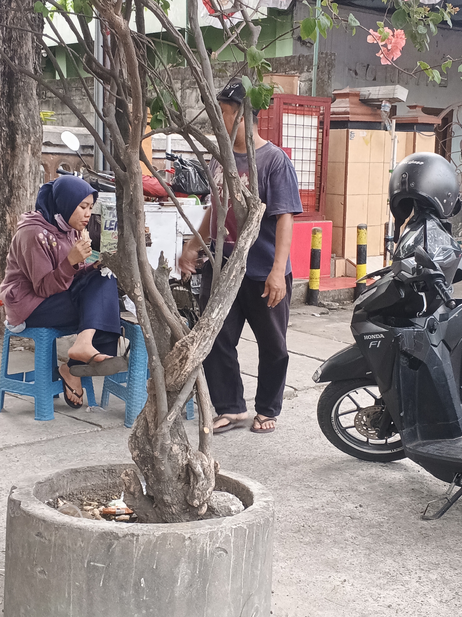 In Krian, Indonesia, a woman wearing a blue hijab and a mauve hoodie sits on a blue plastic stool, eating. She is positioned to the left of a tree in a concrete planter. A man in a dark blue t-shirt and black pants stands to the right of the tree, partially obscuring it. A black Honda motorcycle with a helmet on its seat is parked to the right of the man. The background shows buildings with orange and red accents, and a red gate. The scene appears to be on a sidewalk or street.