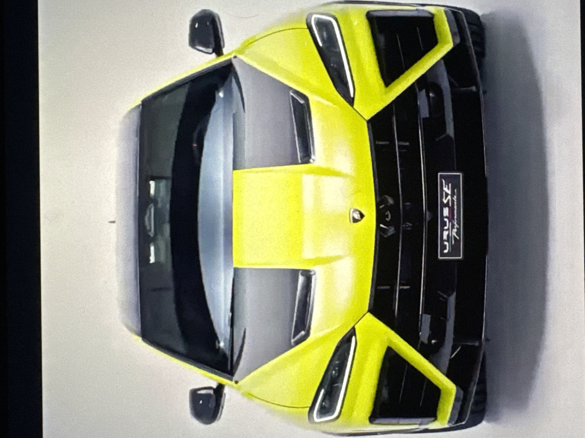 The image displays the front view of a bright yellow Lamborghini Urus SE parked on a white-ish surface, likely a showroom or studio. The vehicle has a black hood with a prominent yellow stripe running down the center, and a black grille with angular, aggressive styling. The headlights are sleek and modern, with distinct LED daytime running lights. A small rectangular badge on the lower grille reads "URUS SE Performance". The background is a plain, light-colored wall, suggesting a controlled environment rather than an outdoor setting in Sant'Angelo di Piove di Sacco, Italy. There are no people or other objects visible in the scene.