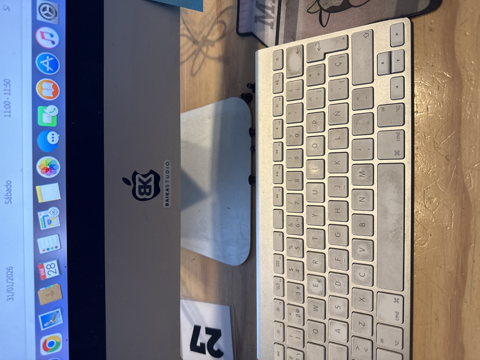 The image depicts an indoor scene featuring a desktop computer setup on a light-colored wooden desk. The central subject is an Apple iMac computer, with its silver-colored lower bezel and stand visible. Affixed to the lower bezel is a black sticker featuring a stylized "BK" logo within an Apple silhouette, and a smaller white rectangular sticker beneath it that reads "BAIKASTUDIO".
The iMac's display shows a macOS desktop environment. The top menu bar indicates the date "31/01/2026", the day "Sábado" (Saturday), and the time "11:00 - 11:50". A dock of application icons is visible, including Chrome, Mail (with a "28" notification on the calendar icon, possibly for ENE 28), Notes, Photos, Safari, and other standard macOS applications.
In front of the iMac, an Apple Magic Keyboard is positioned on the desk. The keyboard exhibits visible wear and dirt accumulation, particularly on the spacebar and several alphanumeric keys. The keyboard layout includes Spanish characters such as 'Ñ' and 'Ç'. Directly below the iMac's stand, several small, dark, cylindrical objects, possibly screws or debris, are scattered on the desk surface. To the left of the iMac stand, a white card or piece of paper with the number "27" printed in black is partially visible. To the right of the keyboard, a segment of a mousepad with a cartoonish cow graphic and the partial letter "M" is visible.
Based on the "BAIKASTUDIO" identifier and the Spanish keyboard layout, the location of this scene is identified as an office or studio in Puebla City, Mexico.