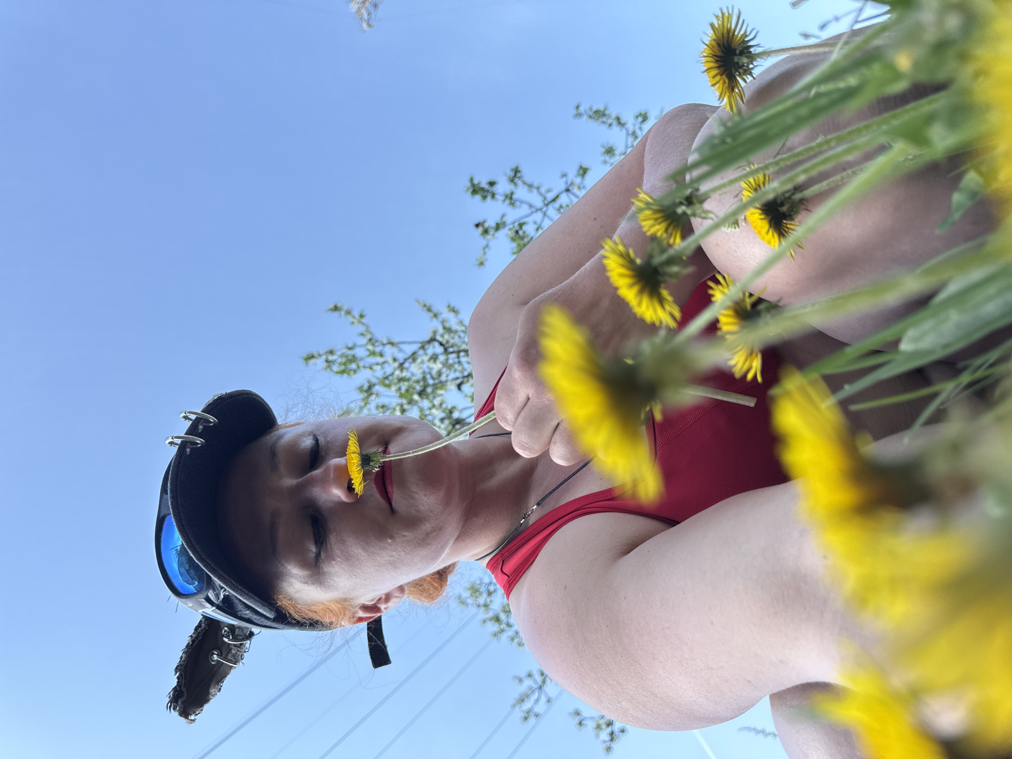 A low-angle, upward-facing medium shot captures a person with fair skin in a red garment holding a dandelion flower to their nose. The person, wearing a dark hat with goggles and a feathered extension, has red lipstick. The background includes a clear, blue sky and sparse tree branches with light green leaves. Yellow dandelions surround the person. The location is not discernible from the image alone.