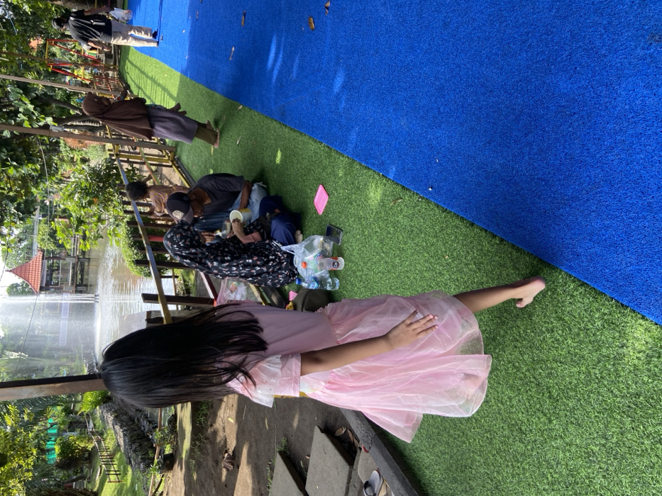 A young girl in a pink dress walks on artificial grass, looking away from the camera. Two adults and a child are seated on the artificial grass to the left, eating. They appear to be picnicking near a fountain in a public park in Kecamatan Sidoarjo, Indonesia, located at coordinates -7.441 latitude and 112.705 longitude. Behind the girl is a wooden fence, and beyond the fence is a pond with a fountain in the middle. There is a small building to the left of the fountain. The park is well-maintained and has a blue artificial grass pathway. A woman in a hijab is walking away from the camera on the pathway to the right. Another man in a red shirt and blue jeans is walking away from the camera in the distance.