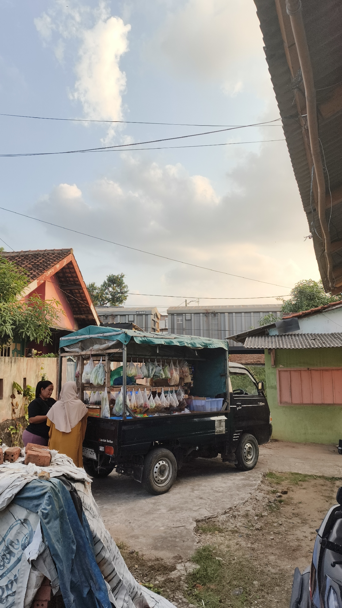 In the city of Bandar Lampung, Indonesia, a small truck is parked and converted into a mobile market stall. The truck, a dark color, has a green tarp covering the top, creating a shaded area for displaying goods. Suspended from the tarp are numerous plastic bags filled with produce. Two women are near the truck, seemingly interacting with the stall. One woman, wearing a black shirt, is standing and looking down. The other woman, in a yellow outfit and a light-colored hijab, appears to be attending to the stall. The background shows a mix of residential structures, including a pink-colored house with a red roof and a green building with a corrugated metal roof. Overhead power lines are visible against a sky with scattered clouds. The scene is likely a residential street or a small market area within the city.