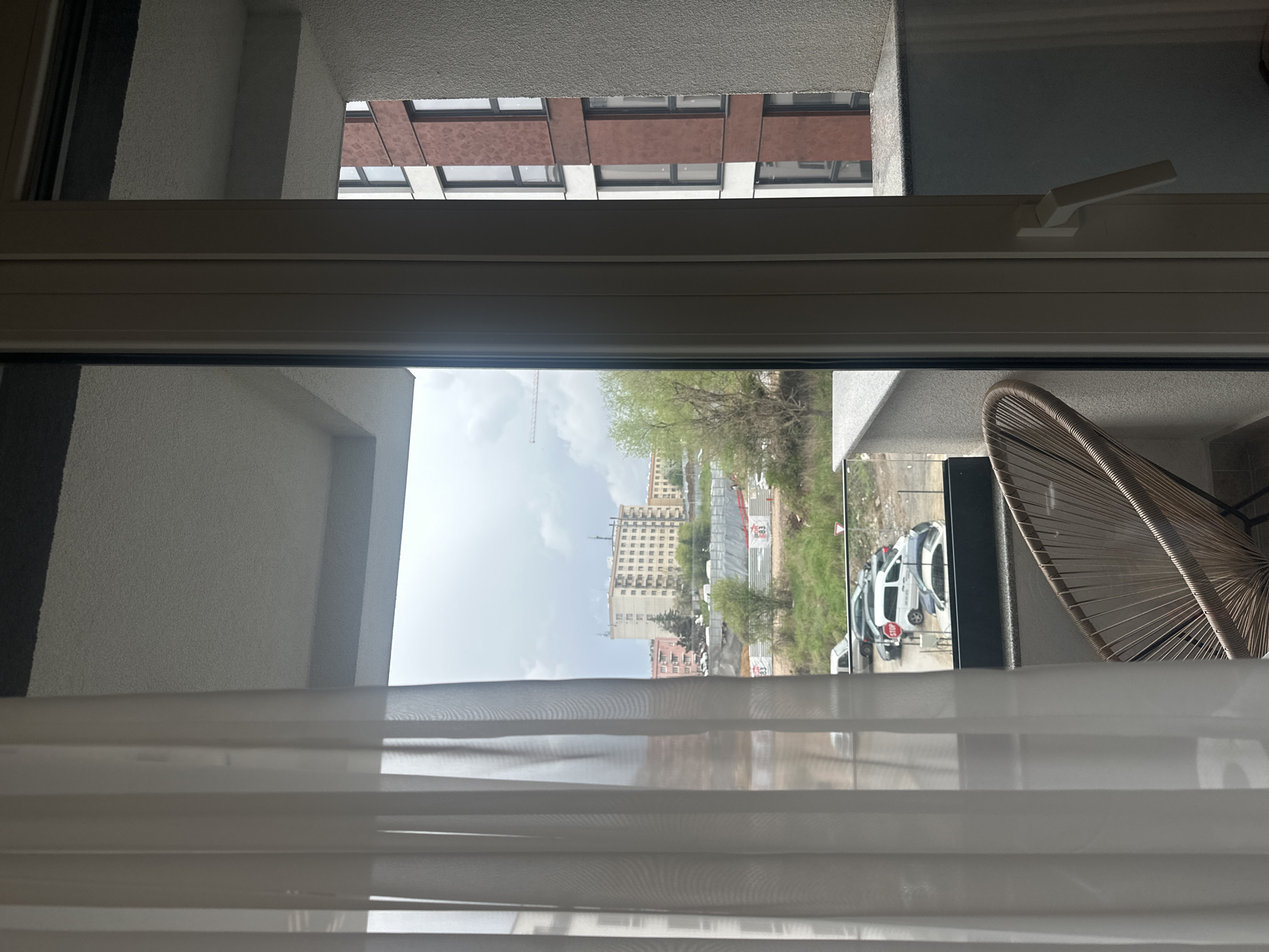 The image, originally captured and displayed rotated 90 degrees clockwise, presents an indoor perspective looking outwards through a window onto an urban scene. A light-colored window frame spans horizontally across the middle of the view, with a white handle visible on the right side.
In the immediate foreground, white, translucent horizontal blinds are partially drawn across the lower section of the window. On the right, just outside the window frame, a segment of a light brown, possibly wicker or rattan, outdoor chair is visible, suggesting a balcony or terrace. The textured, light-colored walls of an adjacent building or balcony are seen above and below the window frame.
Beyond the window, an urban landscape unfolds under an overcast sky, indicating a cloudy daytime. Directly above the main window frame, another building's facade is visible, characterized by a textured light-colored surface interspersed with dark reddish-brown panels and multiple windows.
In the distance, an expansive city view includes several multi-story buildings, notably a large, light-colored structure with numerous windows. Green trees with fresh foliage are scattered throughout the scene, suggesting spring or early summer. Below the distant buildings, a road or open area is visible, where several light-colored vehicles, likely vans or SUVs, are parked. A red "STOP" sign is partially visible on the rear of one vehicle. A grey temporary fence suggests a construction site, and a tall construction crane is prominent against the cloudy sky, hinting at ongoing development. The scene captures a quiet, observational moment of a city environment, consistent with a location like Sofia, Bulgaria. No people are depicted.