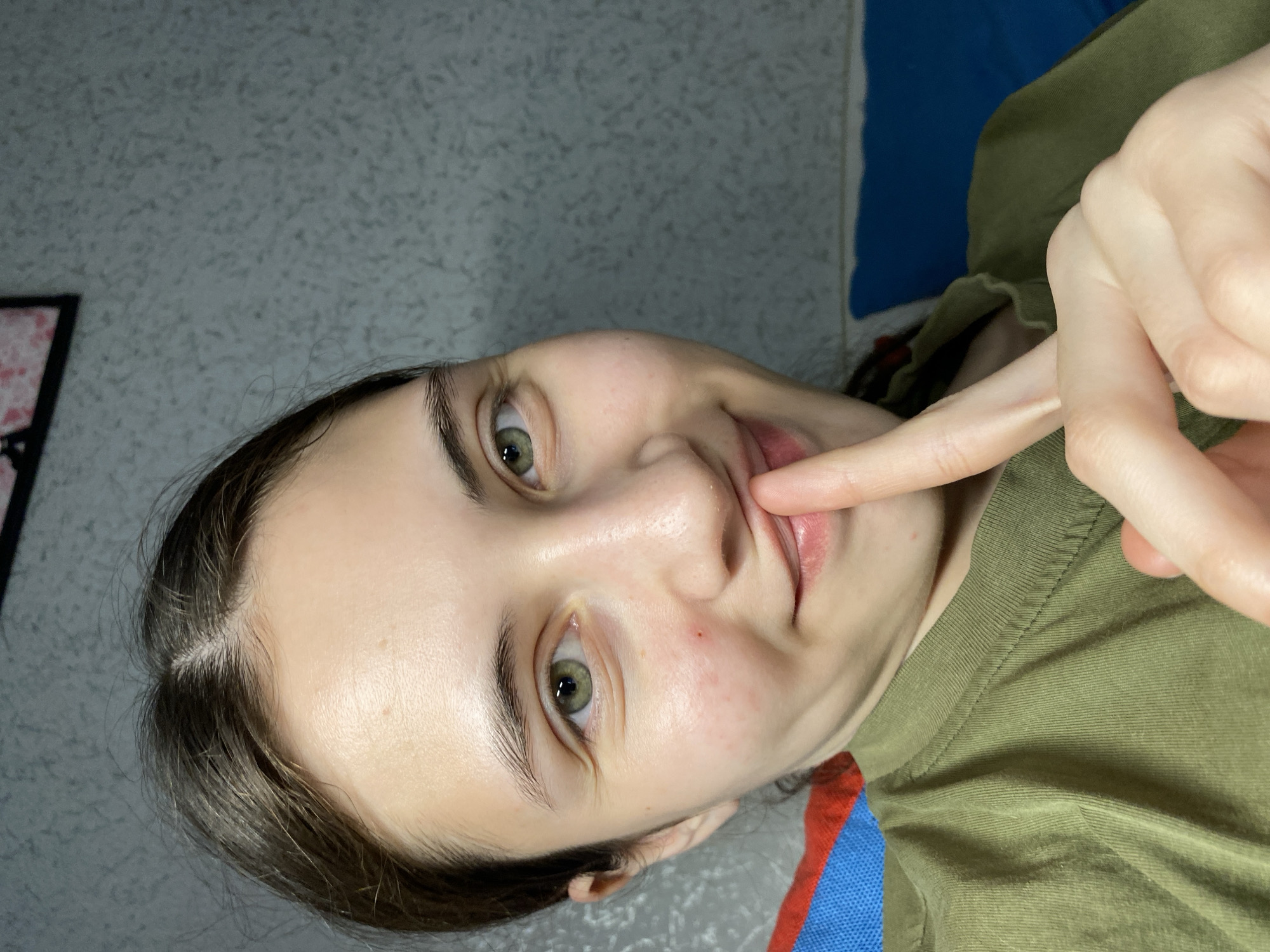 A young woman with fair skin and green eyes is positioned at an angle to the camera. She is wearing a green t-shirt. Her left index finger is extended toward her lower lip. The background is a patterned gray wall with a hint of a blue surface. There is no information to identify the location as Votkinsk, Russia.