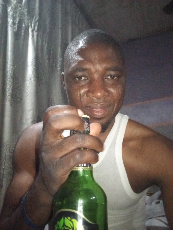 An individual is seen in the photo, holding a green bottle with a black and gold label. The person has dark skin, short hair, and wears a white tank top. The background appears to be a curtain. The person looks directly at the camera with a slight smile. The overall lighting seems dim, highlighting the contours of the individual's face.