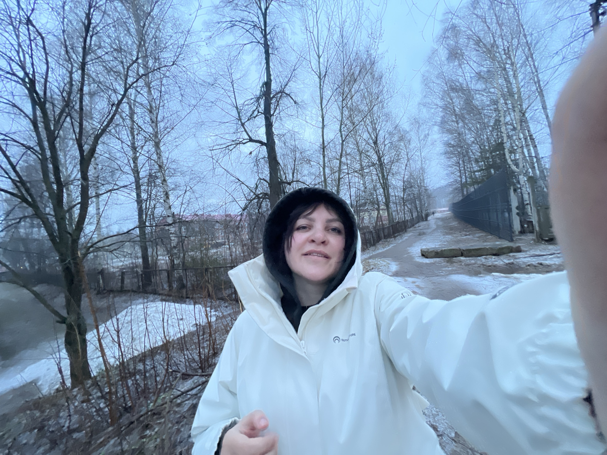 A woman in a white hooded jacket takes a selfie outdoors on a cold, overcast day. Bare trees surround her, with a patch of snow visible in the foreground and along a path that extends into the background. A dark fence runs along one side of the path. Based on the location of Noviy, Russia, the image likely depicts a winter scene.