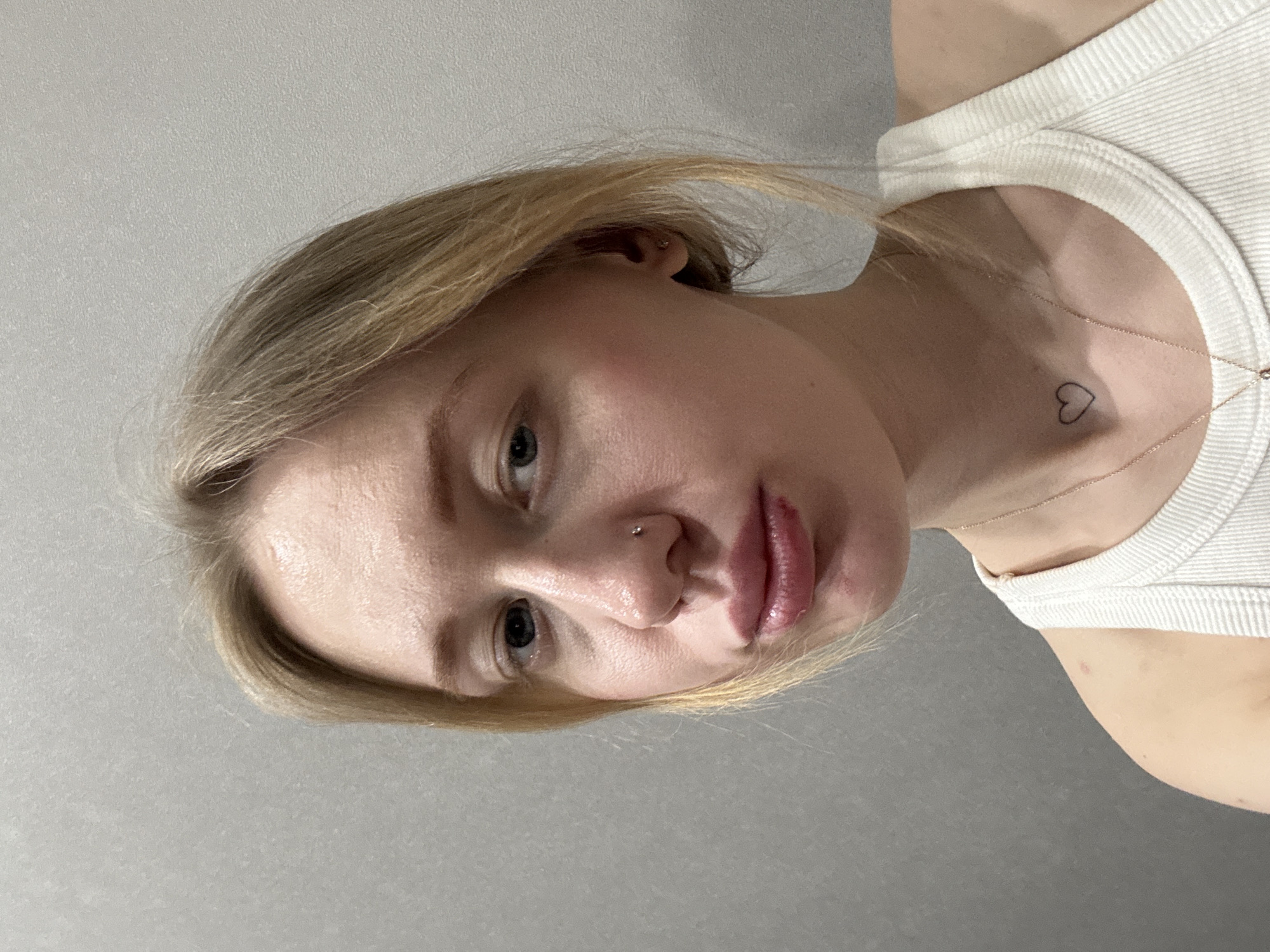 A close-up, chest-up portrait depicts an individual with fair skin and blonde hair, positioned against a plain, light grey, textured wall. The individual wears a white ribbed tank top. A thin gold-colored chain necklace with a small, indistinct pendant is visible, along with a small, black outlined heart tattoo on the upper left chest/collarbone area. A silver stud piercing is present in the left nostril. The eyes are light-colored and directed towards the right side of the frame. The lips are full; the lower lip exhibits a red abrasion on its right side. A small stud earring is visible in the right earlobe. The individual's expression is neutral. The image contains no visual cues identifying a specific geographic location such as Rostov-on-Don, Russia.