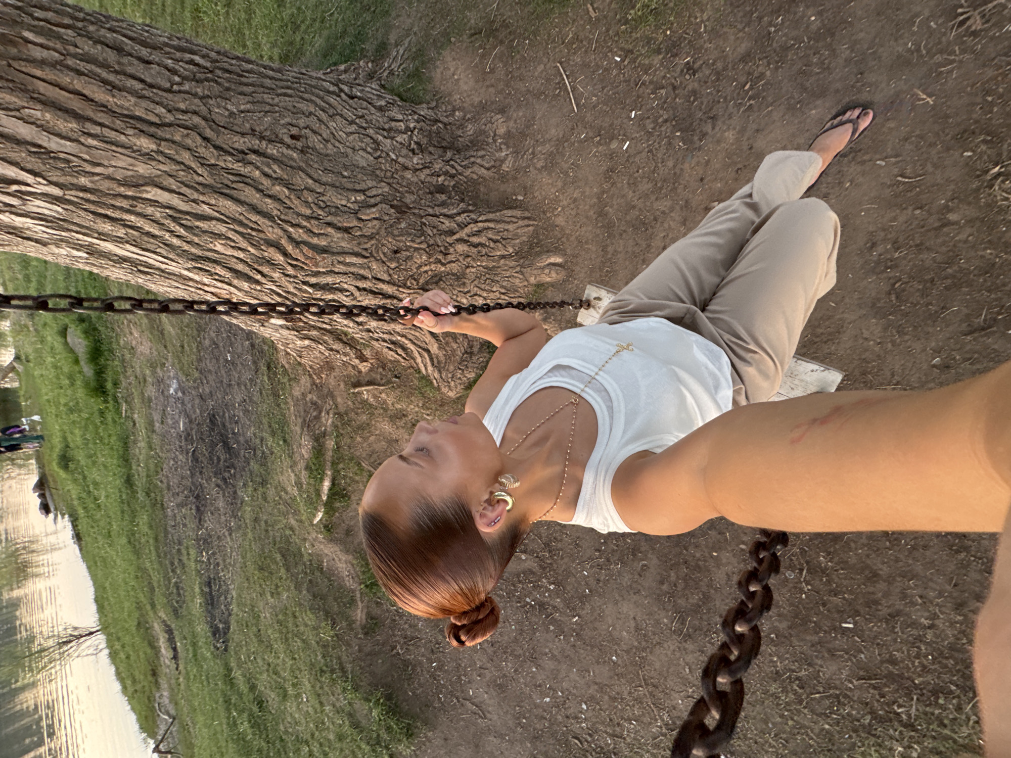 A woman in a white tank top and khaki pants sits on a swing suspended from a tree in Chaikovsky, Russia. She wears sandals and a necklace. The swing's chains are visible. The ground around the tree is dirt and grass, with a body of water and trees in the background. The picture appears to be taken at eye-level.