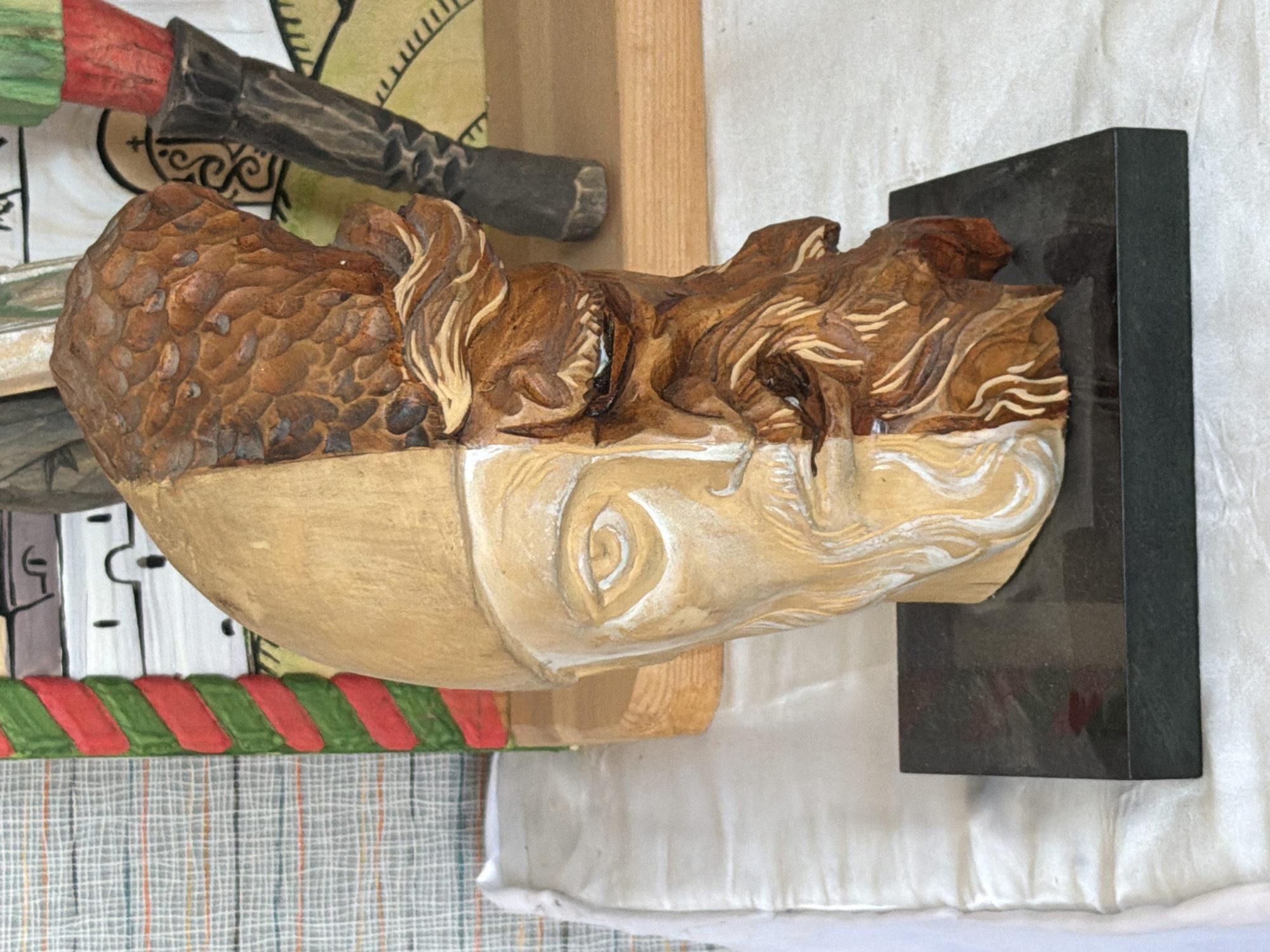 This is a close-up, eye-level shot of a half-finished wooden sculpture of a human head, displayed on a dark, rectangular base. The sculpture is bisected vertically, with the left side being smooth, pale wood with white painted details suggesting a face, and the right side being rough, darker wood with detailed carving representing hair, a beard, and facial features. The backdrop is busy, with a patterned fabric on the left and painted elements suggesting figures or scenery on the right. The sculpture appears to be on a table covered with white fabric. No specific location in Lebedyan, Russia is identifiable from the image.