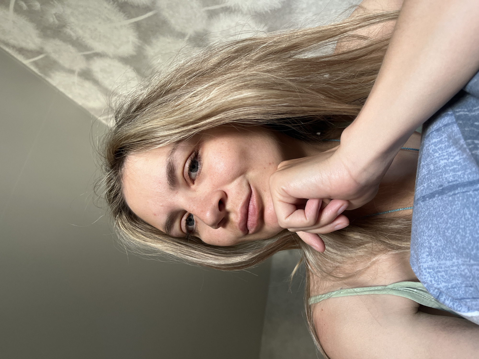 The image shows a blonde woman in a light green tank top and blue blanket. Her hair is parted to the side and her hand is resting on her chin. The background includes a gray wall and wallpaper with a leaf pattern. The location is in the city of Chaikovsky, Russia.