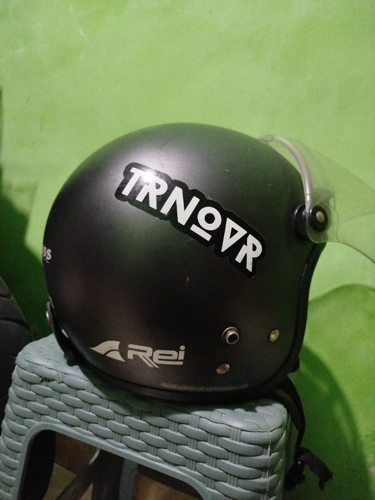 A black, open-face motorcycle helmet with a clear visor sits on a textured, light green plastic stool. The helmet has two white stickers on its side. The upper sticker features the text "TRNOVVR" in a stylized font with geometric elements. Below this, there is a logo that appears to be an abstract bird or wing shape followed by the text "Rei". The background is a textured, light green wall. The overall scene appears to be an indoor setting, possibly a room or a storage area, in Surabaya, Indonesia.