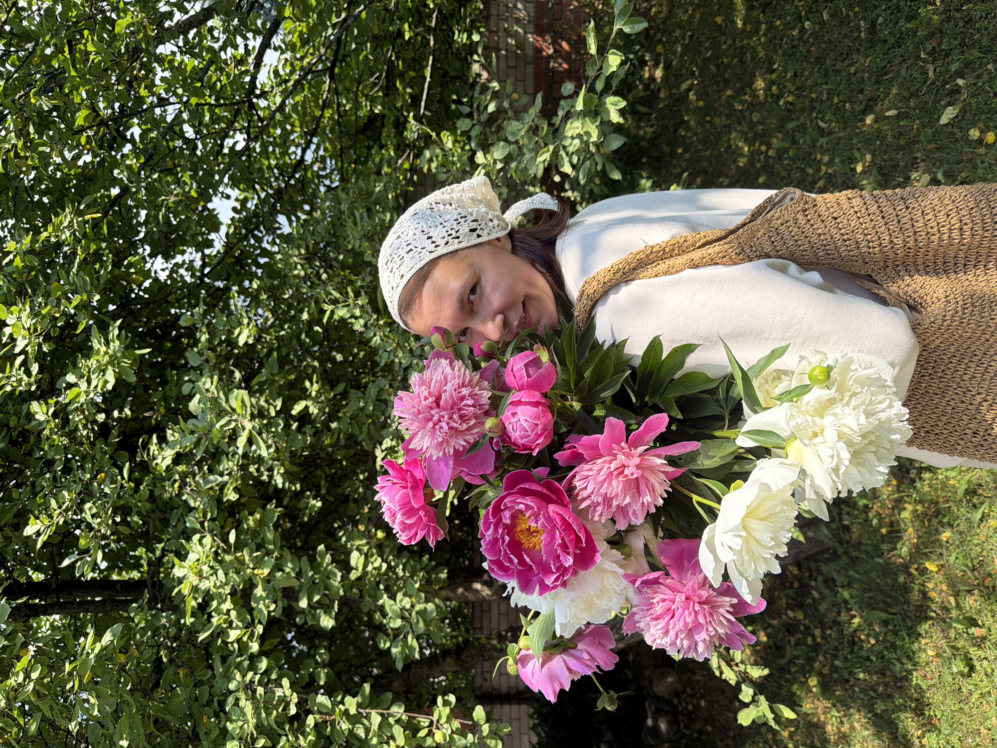A woman with dark hair, wearing a white crocheted headscarf and a cream-colored sweatshirt, stands outdoors holding a large bouquet of pink and white peonies. She is looking towards the camera and smiling. The bouquet is positioned in front of her, obscuring part of her body. She is carrying a woven, tan-colored bag over her right shoulder. The background consists of lush green foliage from trees and a hint of a brick structure on the right. The ground is covered in green grass with scattered fallen yellow leaves. The scene appears to be a garden or yard, likely in a rural or suburban setting, possibly within the Прикамский district of Russia, given the context.