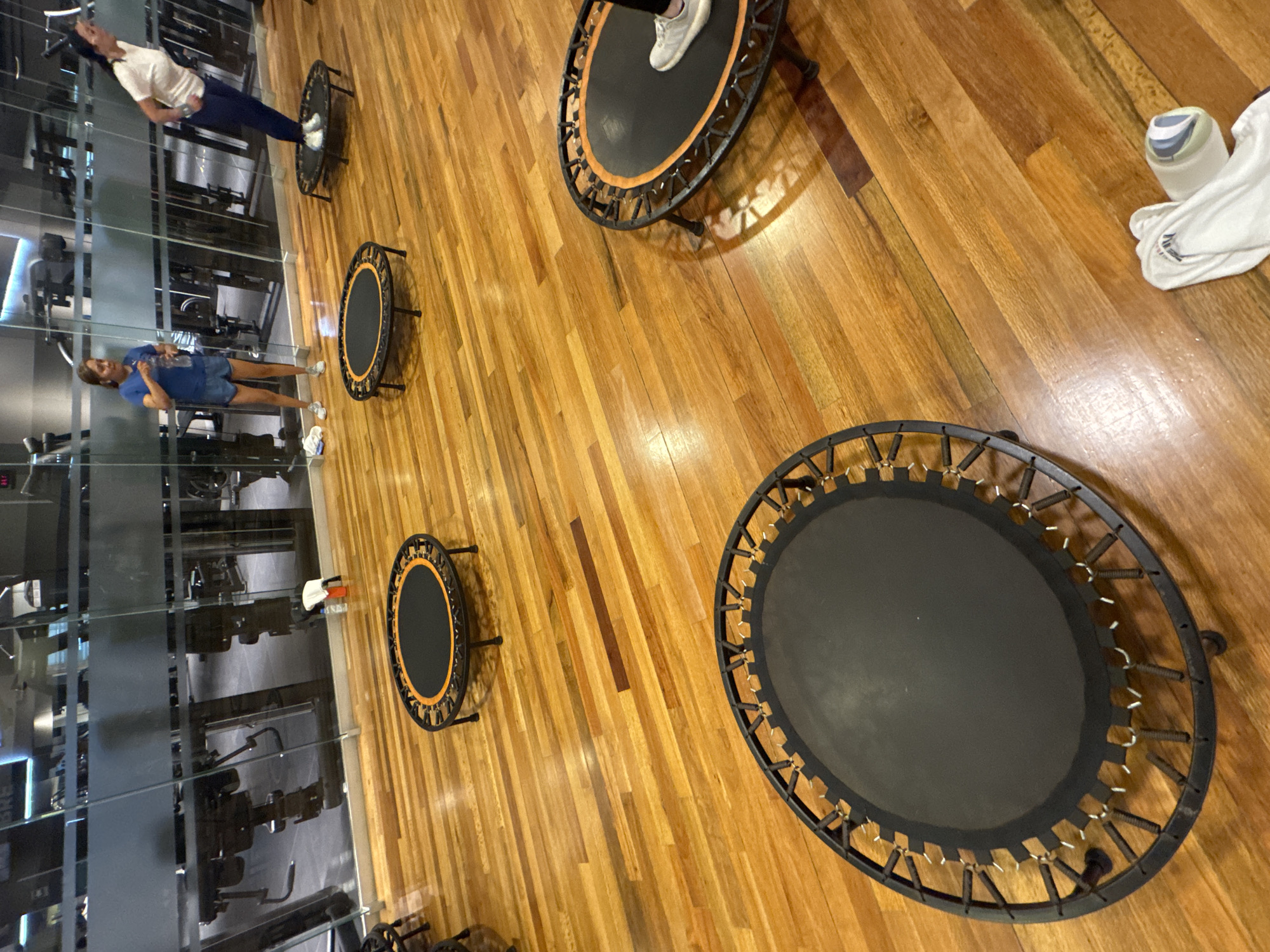 This image captures an indoor fitness studio in Puebla, Mexico, depicting a group exercise setting featuring mini-trampolines. The floor is made of polished light brown and orange-toned wooden planks.
Two women are visible within the frame. One woman, wearing a white long-sleeved top and dark pants, is standing on a black mini-trampoline (rebounder) with an orange ring and black springs. Her right foot, clad in a white sneaker, is clearly positioned on the trampoline mat. She appears to be in the middle of an exercise or balancing, facing away and slightly to the left of the camera's perspective. Another woman, dressed in a blue t-shirt and blue shorts, is standing on the wooden floor to the left, near another black mini-trampoline. She holds a transparent water bottle in her right hand and appears to be looking towards the left.
Several black mini-trampolines are arranged across the wooden floor, each featuring a dark mat and either orange or white-colored elastic bands or springs connecting the mat to the frame. A white towel with blue stripes is casually placed on the floor near one of the trampolines.
The background features a large glass partition, reflecting various pieces of gym equipment such as weights, benches, and exercise machines, suggesting another area of the fitness facility. Due to the reflections and angle, some indistinct text, possibly "BRE", can be partially observed on the glass. The scene conveys a well-lit, active indoor environment, likely during a fitness class or training session.