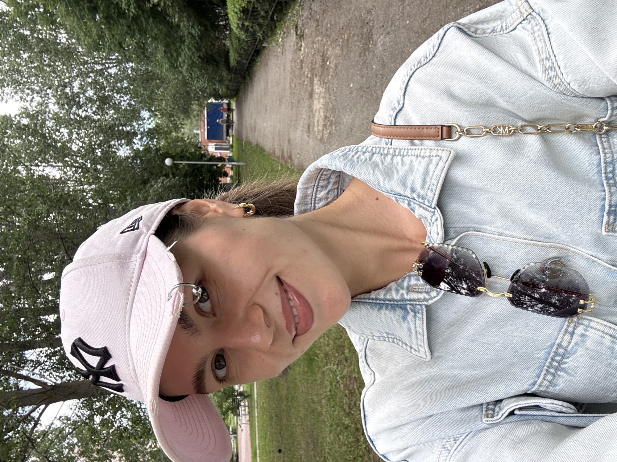 A woman is photographed outdoors, in a medium close-up selfie. She is wearing a light pink baseball cap with a black logo, a light blue denim jacket, and a gold hoop earring. Sunglasses with dark lenses and gold frames are hanging from the neckline of her jacket. A brown leather purse strap with a gold chain detail is visible over her right shoulder. She has a mole on her neck and a small ring through her eyebrow. The background consists of a paved walkway lined with trees and green grass. In the distance, two people are walking away from the camera, and a blue truck is parked. The overall lighting suggests an overcast day.