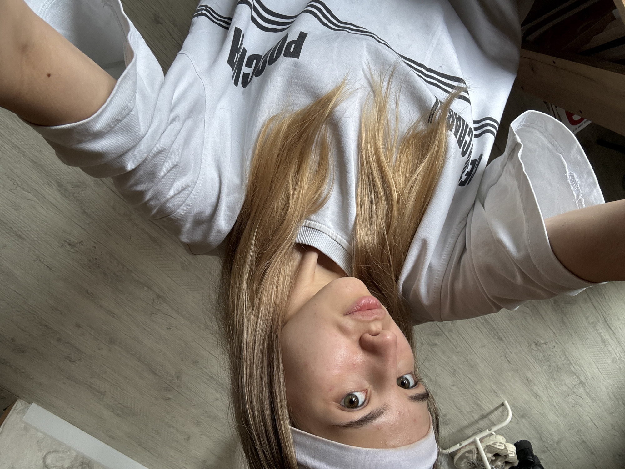 A young, light-skinned woman with blonde hair and brown eyes is lying on a light gray, wood-look floor. She is wearing a white T-shirt with black lettering on the front and a light purple headband. The photograph appears to be taken from her perspective, looking down at herself. In the background, there are items under what looks like a wooden bed frame. The location is unidentified; the text request to identify the location in the city of Noviy, Russia, appears irrelevant.