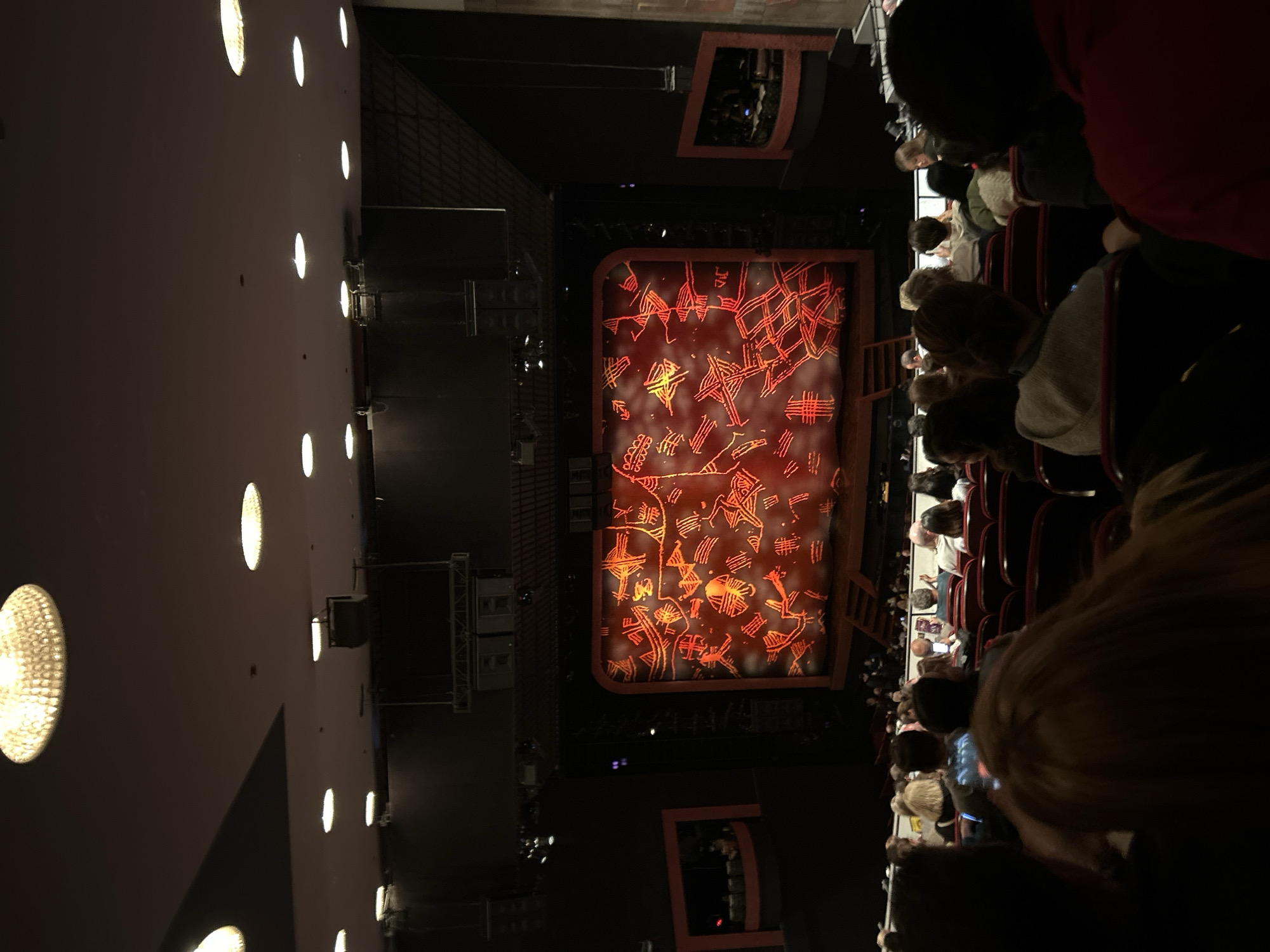 The image shows a theater performance viewed from a high angle, likely from a balcony or upper level. The stage is set with a large backdrop featuring a reddish-brown design resembling ancient glyphs or tribal art. A few rows of audience members are visible in the foreground and along the right side of the image. They appear to be seated and facing the stage. The theater has a dark interior with dim lighting, except for the stage which is illuminated. A wall with a series of circular lights runs along the left side of the image. Additional structural elements like lighting rigs are visible above the stage. The specific location within New York City is impossible to ascertain with only this image.