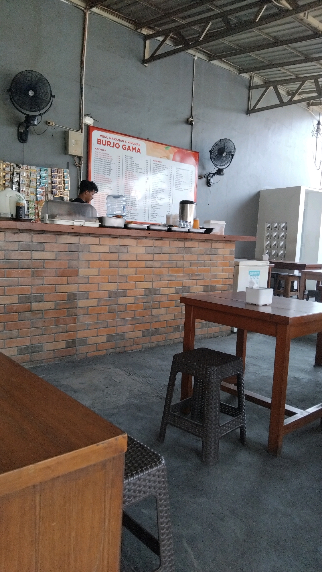 This is an indoor scene at what appears to be a food stall or eatery, likely located in Proyonangg, Indonesia, given the context provided. The foreground features a wooden table with a woven plastic stool tucked underneath it. To the left, another wooden surface, possibly a counter, is visible. In the midground, a brick counter spans the width of the image, behind which a young male employee is attending to customers. Above the counter, a large menu board displays food and drink items, with "BURJO GAMA" prominently displayed at the top. Various items are arranged on the counter, including what appears to be a water dispenser and trays of food. The background shows a grey wall with industrial-style roofing and ventilation fans. Further back, tables and chairs are set up for dining, and a white appliance, possibly a refrigerator, is present. The flooring is a dark concrete. The lighting suggests it is daytime, possibly with artificial lighting supplementing natural light.