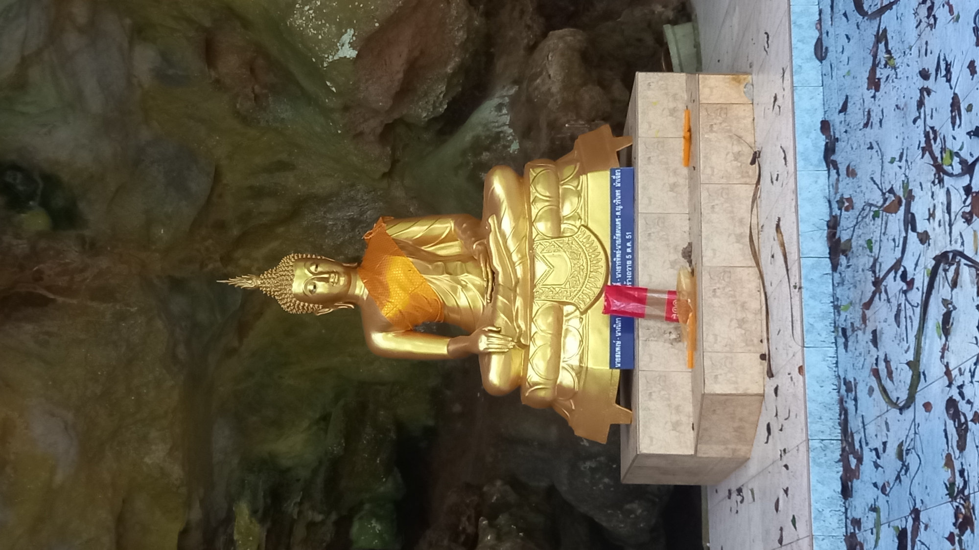A gilded statue of Buddha sits in meditation on a multi-tiered beige tile base. The statue wears a yellow cloth draped over its shoulder. A yellow candle is placed on the step to the statue's right. Red-and-white striped tape and signage written in Thai are on the step in front of the statue. The statue is placed against a backdrop of mottled rock formations. Dead leaves litter the concrete floor. The city of Ban Pak Ra is not visible in the image.