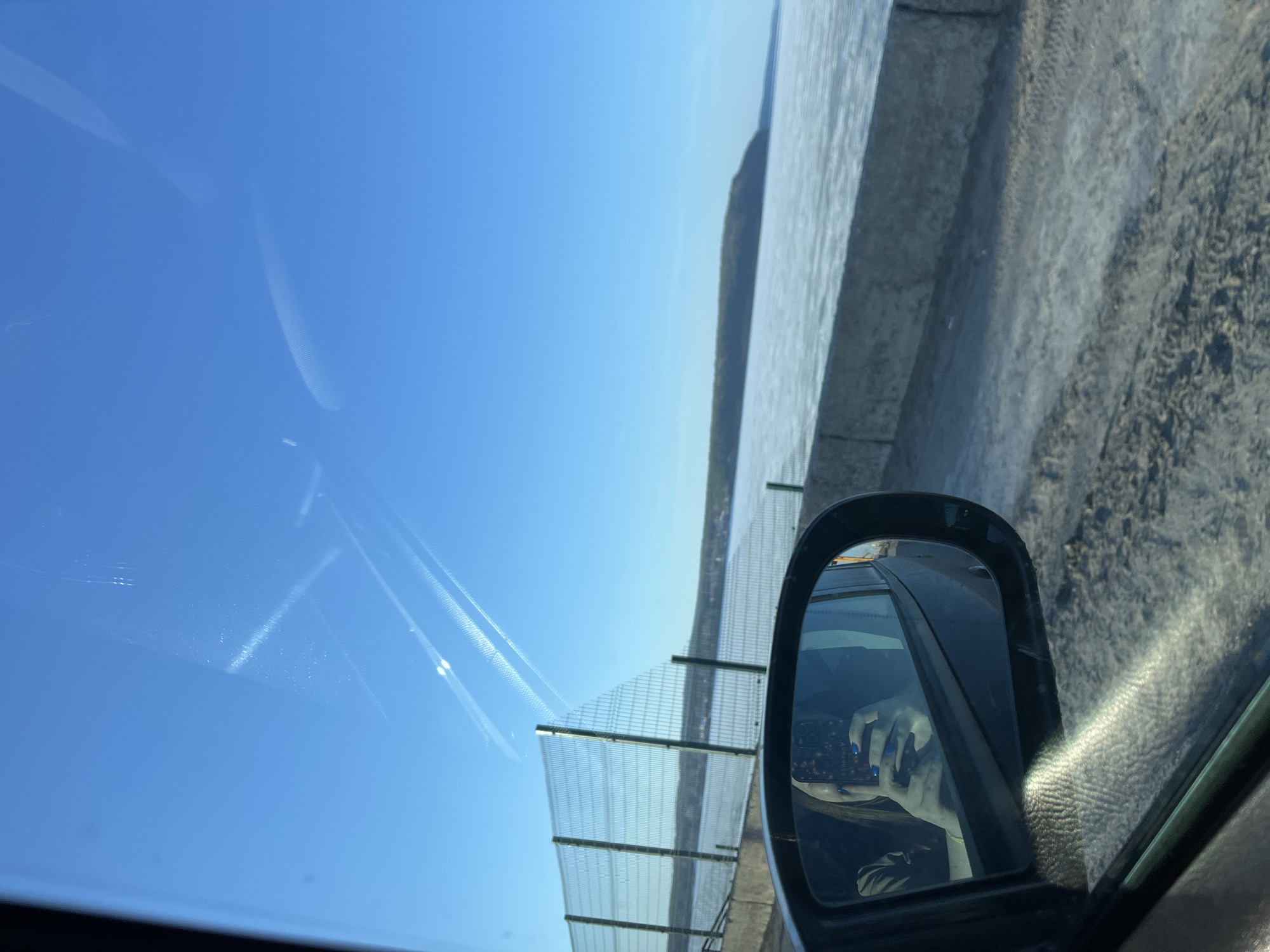 The image is taken from inside a car, likely on the move. The window on the left offers a view of a bright blue sky with contrails. Outside the car, there is a metal barrier, possibly along a road or bridge. Beyond the barrier, a body of water can be seen under the same bright sky. The car's side mirror is visible in the foreground, reflecting the interior of the car, including a hand holding a phone with a decorated case. Given the prompt, it is possible this scene is within the city of Chaikovsky, Russia, though the provided image does not contain any indicators to definitively confirm the location.