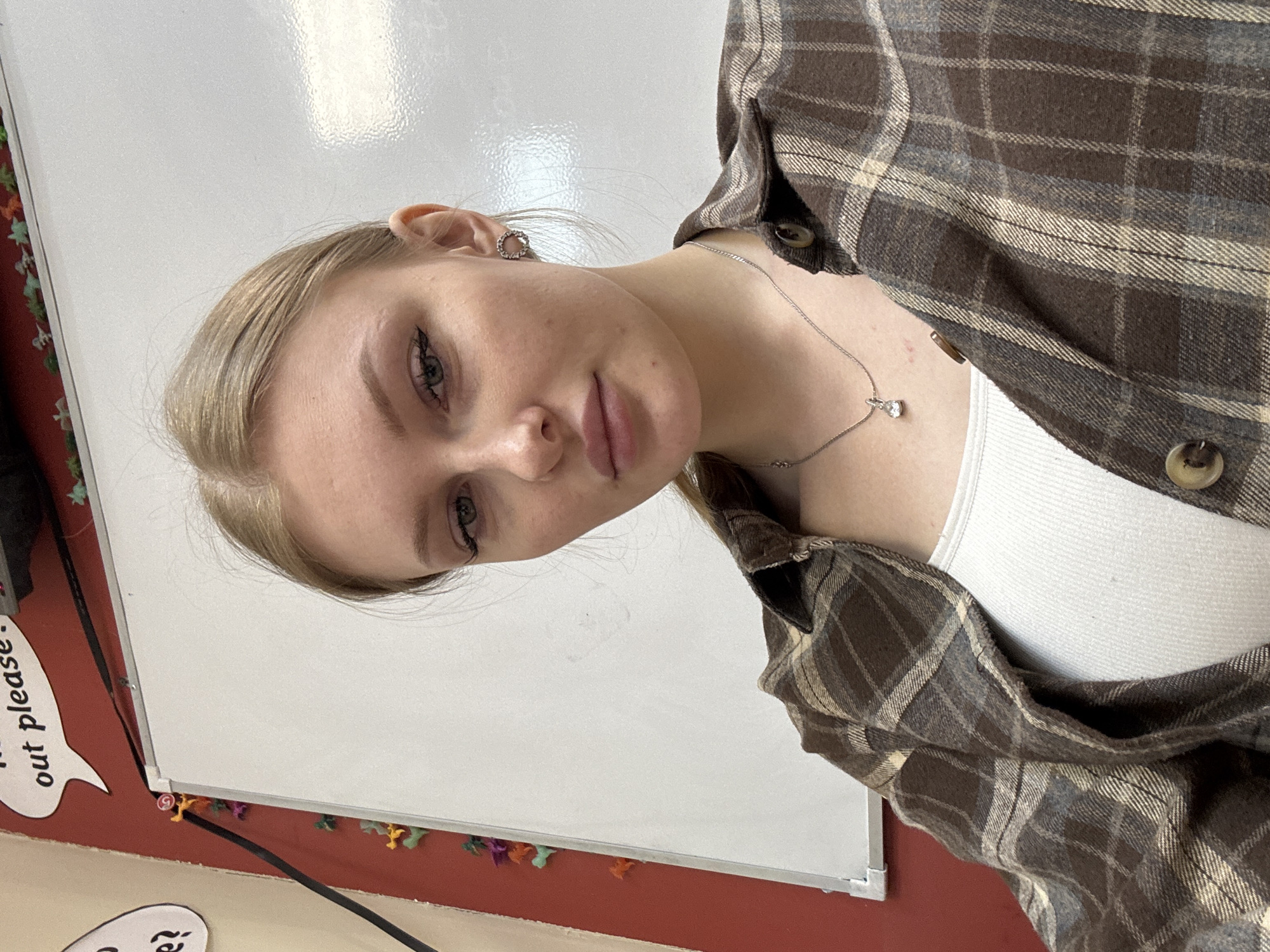 A young woman with blonde hair and light skin is posing for a selfie in an indoor environment. She is wearing a white ribbed tank top and a brown plaid flannel shirt. She also wears a silver necklace with a small pendant and earrings. The background includes a white board with some objects attached to the bottom and left side of the frame. Location: Not detectable.