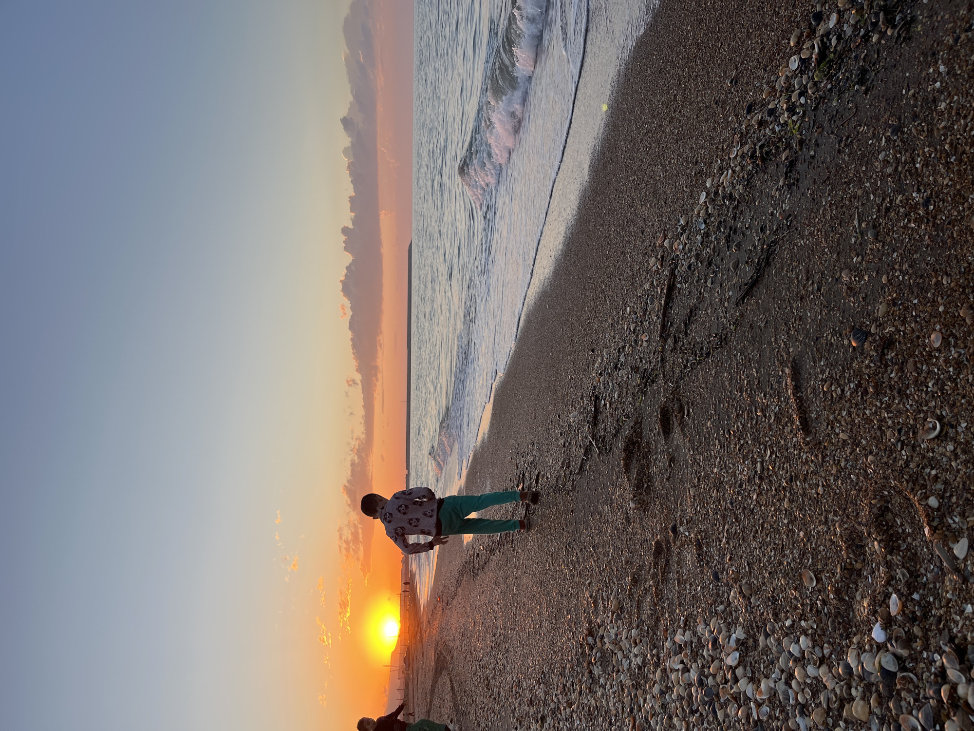 A person stands on a beach at sunset in Kaspiysk, Russia. The beach is composed of dark sand and numerous shells. To the person's right, the ocean laps at the shore with a small wave. The sunset casts an orange glow across the sky, with a visible sun near the horizon. The person is wearing a patterned shirt and green pants.
