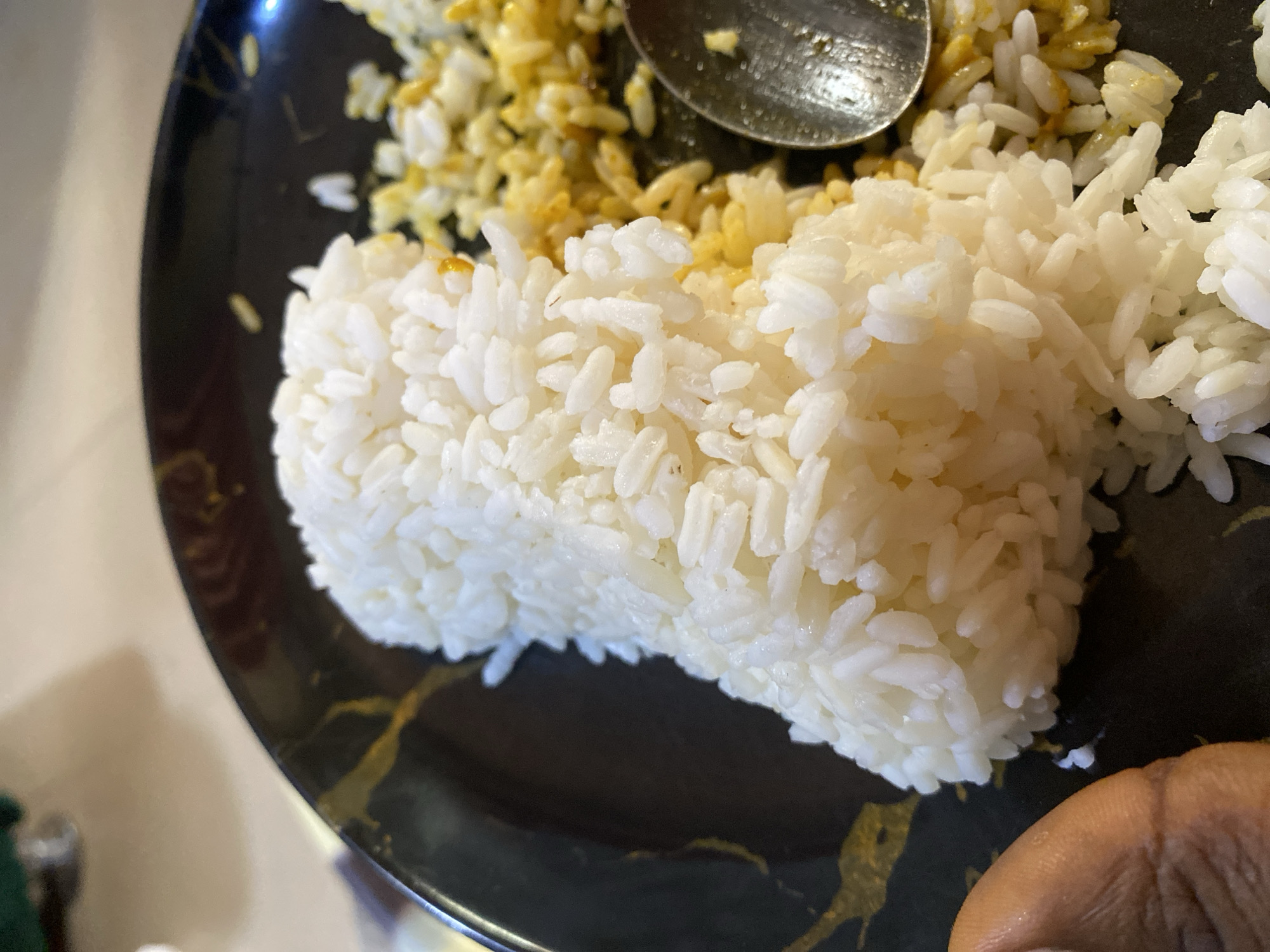 On a black plate, two kinds of rice are visible, white rice molded into a rectangle and yellow rice scattered next to it. A metal spoon rests on the yellow rice. A dark-skinned hand is visible in the lower right corner of the image. The image does not contain any information about the city of Nsukka, Nigeria.