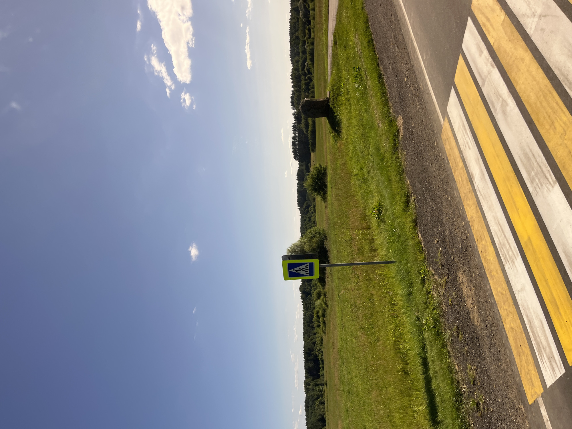 The image depicts a rural roadside scene on a sunny day in Юнькаўскі сельскі Савет, Belarus. In the foreground, a crosswalk with yellow and white stripes is visible on a paved road. To the left of the road, a pedestrian crossing sign is planted in a grassy field. The field extends into the distance, with a line of trees forming the horizon. A large, solitary rock stands in the field to the right of the road, casting a shadow. The sky is a clear blue with a few scattered white clouds. The overall impression is one of a quiet, open landscape.