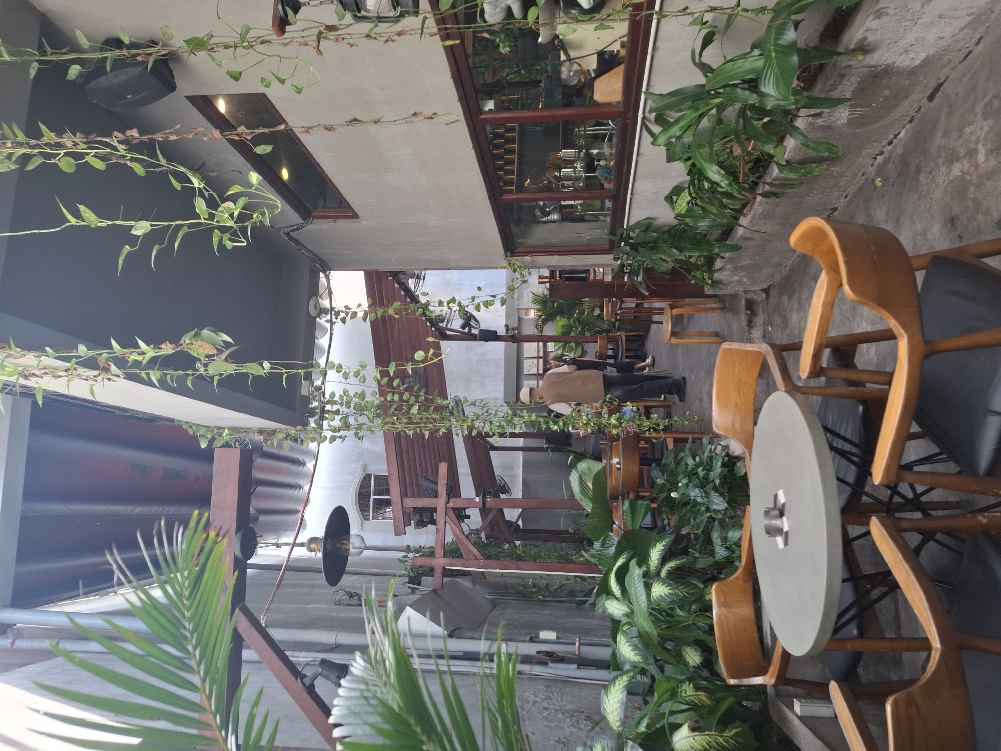 The image depicts an outdoor seating area, likely a cafe or restaurant, in Nha Trang, Vietnam. The scene is characterized by a mix of industrial and natural elements. A concrete floor and grey walls are juxtaposed with a wooden pergola structure overhead, from which hanging plants cascade down. Several wooden chairs with black seats surround an oval-shaped, grey table in the foreground. An ashtray with two cigarette butts rests on the table. In the middle ground, a man wearing a hat and a vest is visible, walking past a seating area. Beyond him, through a large window with a wooden frame, shelves stocked with various items are discernible inside the establishment. The overall ambiance suggests a casual, possibly bohemian, dining or relaxation space.