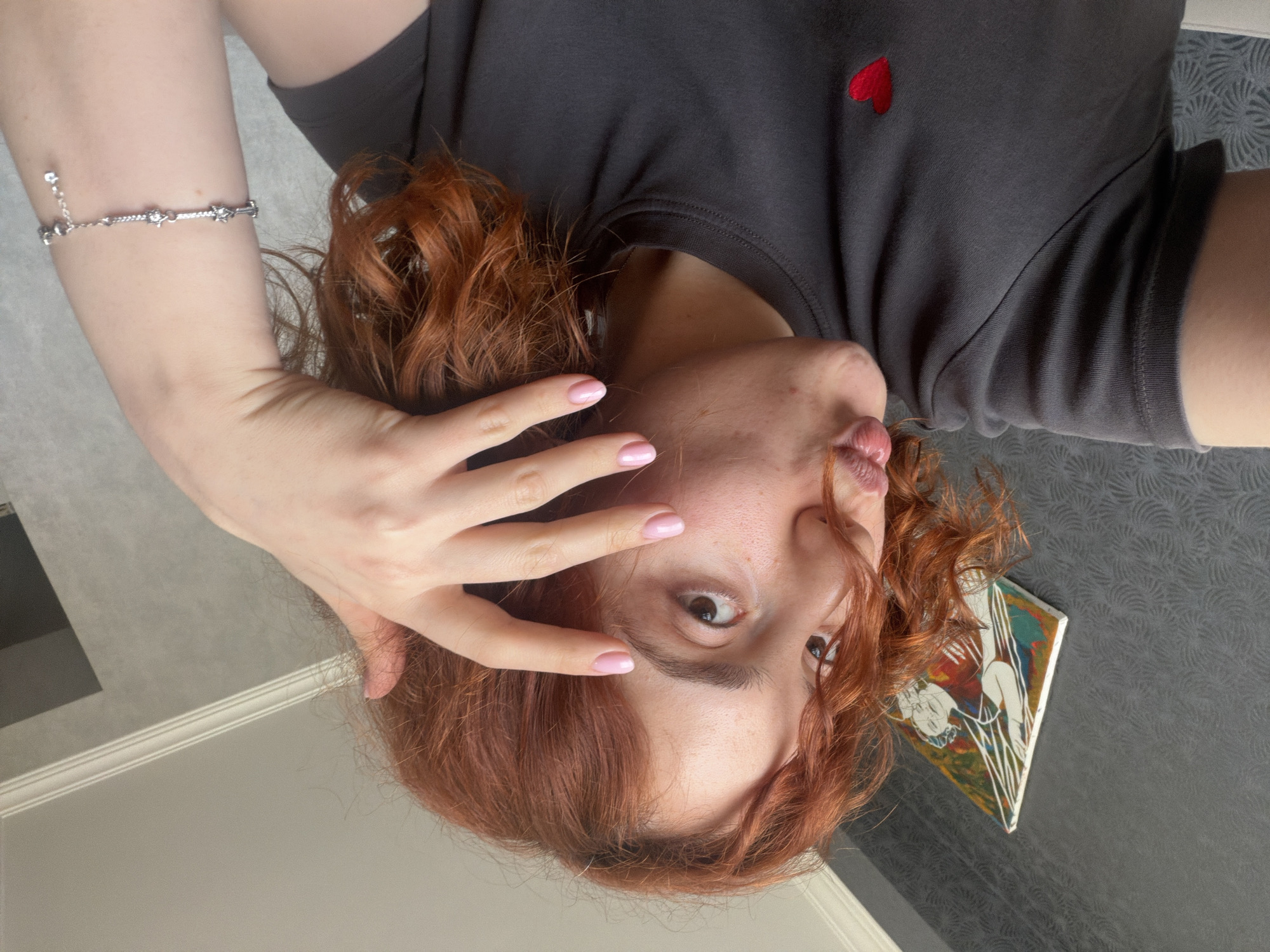 A woman with light skin, red-auburn curly hair, and pink nails is angled downward towards the camera. She is wearing a gray t-shirt with a small red heart emblem on the upper right side. Her left hand is raised with her fingers spread near her eye, and she is pursing her lips. She wears a silver bracelet on her left arm. The background consists of a gray patterned wall and a white wall, along with a small diamond-shaped artwork depicting a white figure on a colored background. The location is not identifiable from the provided image, and the city of Tbilisi, Georgia cannot be confirmed.