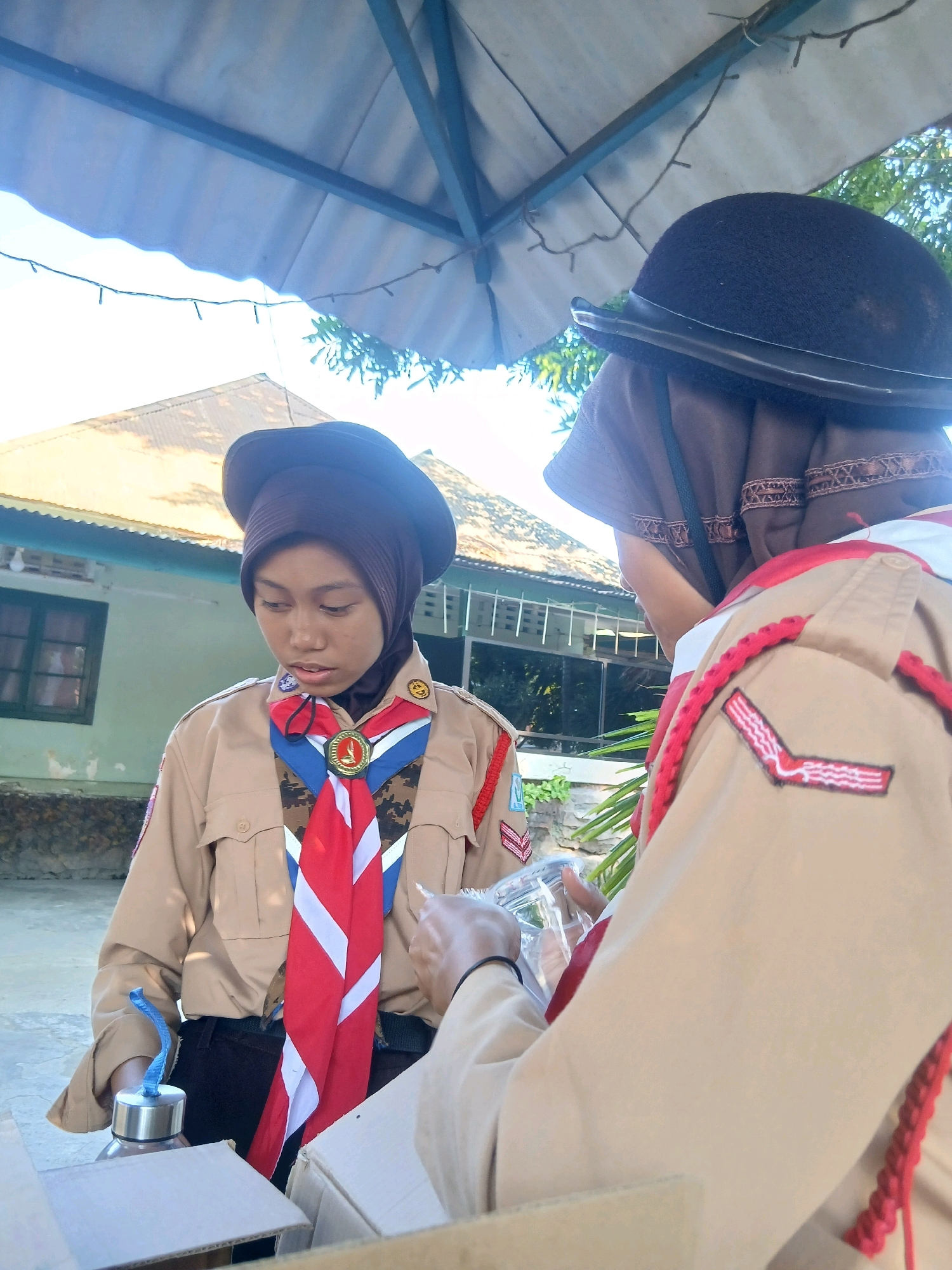 Two young Indonesian girls in scout uniforms are positioned outdoors under a covered structure. The girl on the left, facing the camera, wears a brown hijab and a brown scout hat. Her uniform is a tan short-sleeved shirt with red and white insignia on the collar and a red, white, and blue neckerchief. A gold medallion is pinned to her neckerchief. She is holding a metal water bottle with a blue strap. The girl on the right, seen from the back, wears a dark brown hijab and a dark brown hat, partially obscuring her face. Her uniform is a tan short-sleeved shirt with red trim and insignia on the shoulder. Both girls appear to be interacting with an open cardboard box situated in the foreground. In the background, a light-colored building with green trim is visible, along with some foliage and a light blue sky. The scene appears to be in a residential area.