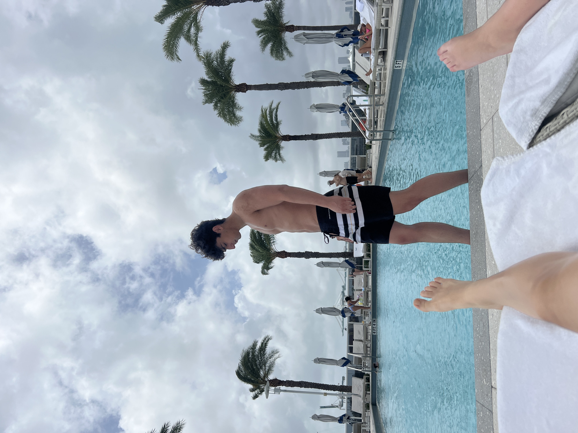The image shows a man in black and white striped swim trunks standing at the edge of a swimming pool. The pool is lined with lounge chairs, some with people sitting on them. Palm trees are visible in the background, and in the distance, a cityscape can be seen. The scene appears to be a rooftop pool deck, likely in Miami Beach, United States of America. The image also shows a foot of a person lying down in a white towel on a lounge chair.