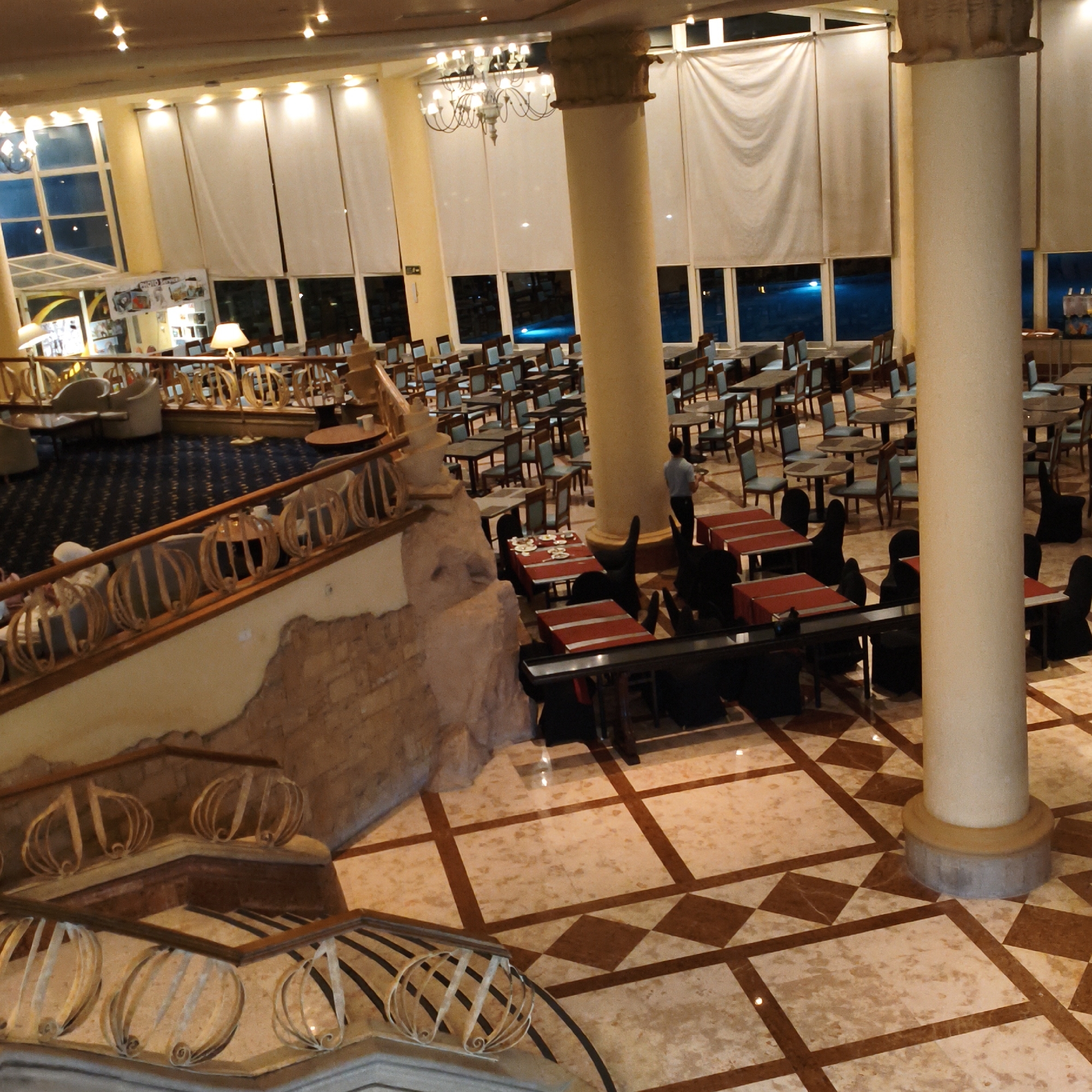 An interior shot captures a grand, multi-level dining area with numerous tables and chairs. The room is illuminated by ceiling lights and decorative chandeliers. Large windows with sheer white curtains line the back wall, offering a view of a dimly lit outdoor area, possibly a pool, with blue lights. The flooring is a patterned marble with squares and diamonds. In the foreground, a wide staircase with decorative railings leads down. A large, cream-colored pillar stands prominently on the right side of the image. A server in a blue shirt and dark pants is seen walking through the dining area with a tray. The overall atmosphere is elegant and spacious, suggesting a hotel lobby or a large restaurant.