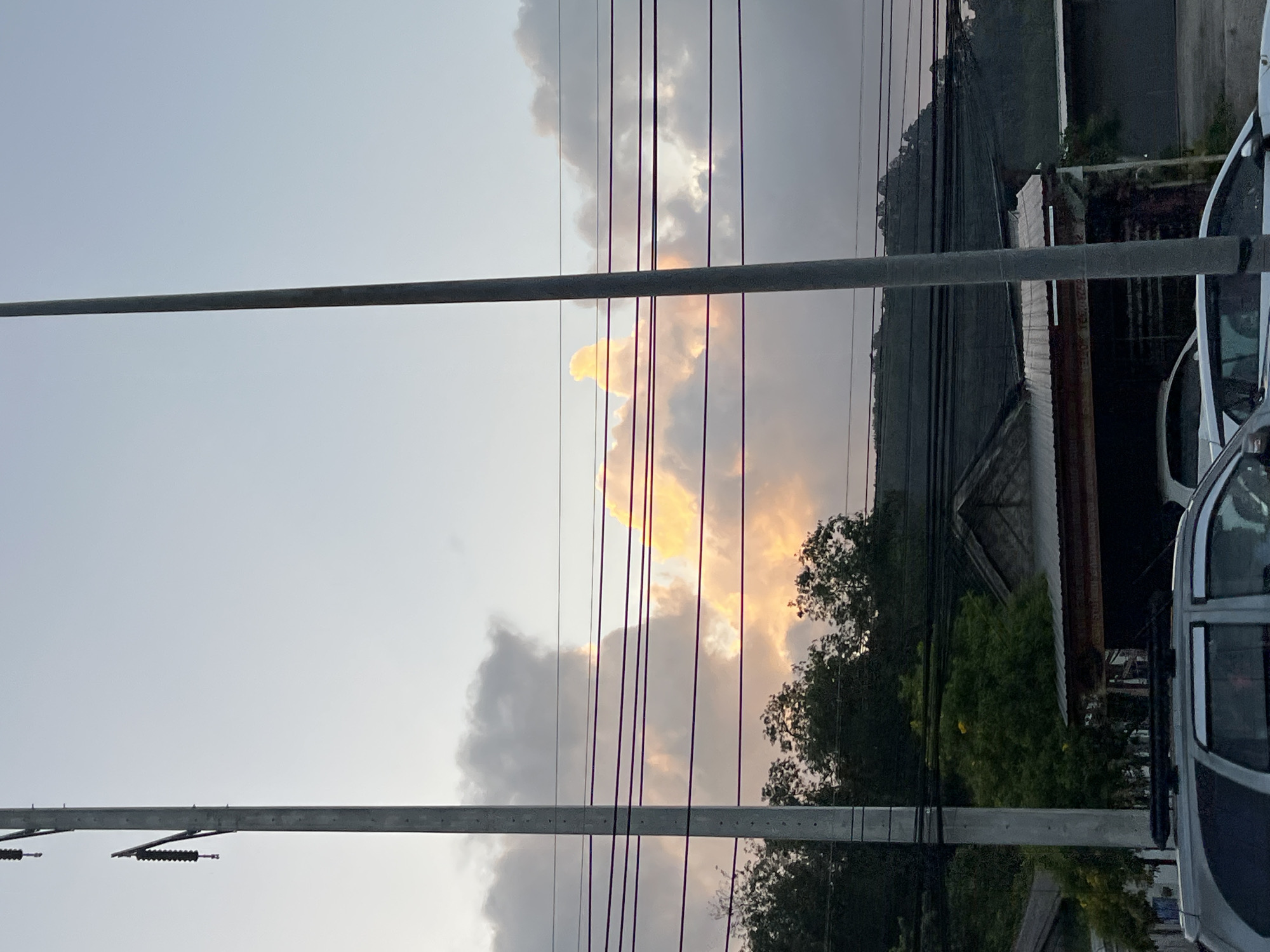 The image captures a view of the sky with dramatic clouds illuminated by the setting sun, casting a warm orange glow. The scene is framed by utility poles and power lines that stretch across the frame, suggesting an urban or semi-urban setting. In the foreground, the roof and hood of a silver SUV are visible, indicating a street-level perspective. Behind the power lines and trees, buildings with corrugated metal roofs are partially visible, and a lush green hillside forms the backdrop. The presence of these elements in Kathu, Thailand, implies a typical residential or commercial area within the district, potentially during the late afternoon or early evening as indicated by the lighting.