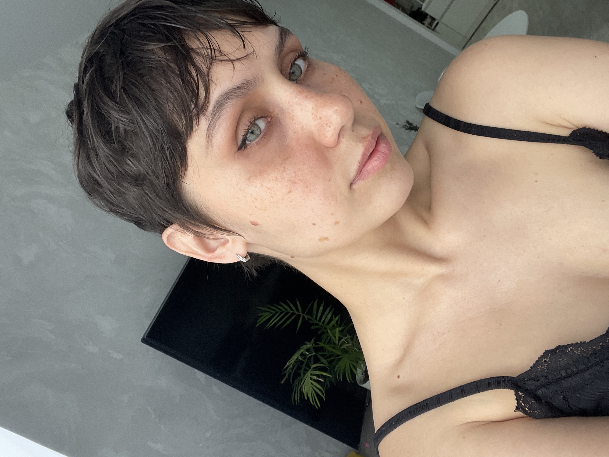 A fair-skinned young woman with short, dark hair and green eyes is the focus of this image. She has freckles and is wearing a black camisole. A plant can be seen in the background, partially obscured by a black object, possibly a TV screen. The setting appears to be an indoor residential space, given the presence of a wall and furniture elements, like the white object. The image is taken at an angle, creating a dynamic perspective. There is no information to suggest that the photo was taken in Izhevsk, Russia.