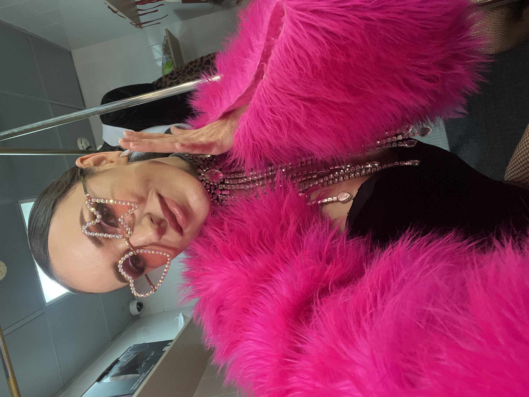 A woman poses for a selfie indoors, looking towards the camera with a subtle smile. Her dark hair is pulled back, and she wears oversized, heart-shaped sunglasses adorned with crystals, featuring pink-tinted lenses. She is clad in a vibrant, fuchsia pink fluffy faux fur coat or boa, a black top underneath, and fishnet stockings visible on her right leg. An elaborate choker necklace with multiple strands of clear crystals and larger central stones dangles long crystal elements down her chest. Her left hand is raised near her face, palm partially visible, fingers slightly spread.
The background suggests an utilitarian indoor space, possibly a dressing room or backstage area. The ceiling is white with a rectangular fluorescent light fixture. White walls are visible, with a small white security camera mounted on the left. A shelf or counter on the left displays a black electronic device box and other items, with a white basin partially visible below. A tall, metallic pole or stand is prominent on the right, reflecting light. Behind it, a dark garment and leopard print fabric are partially visible, along with another shelf holding various containers. The floor is dark. The specific location within the city of Kazan, Russia, is not discernible from the visual information provided in the image.