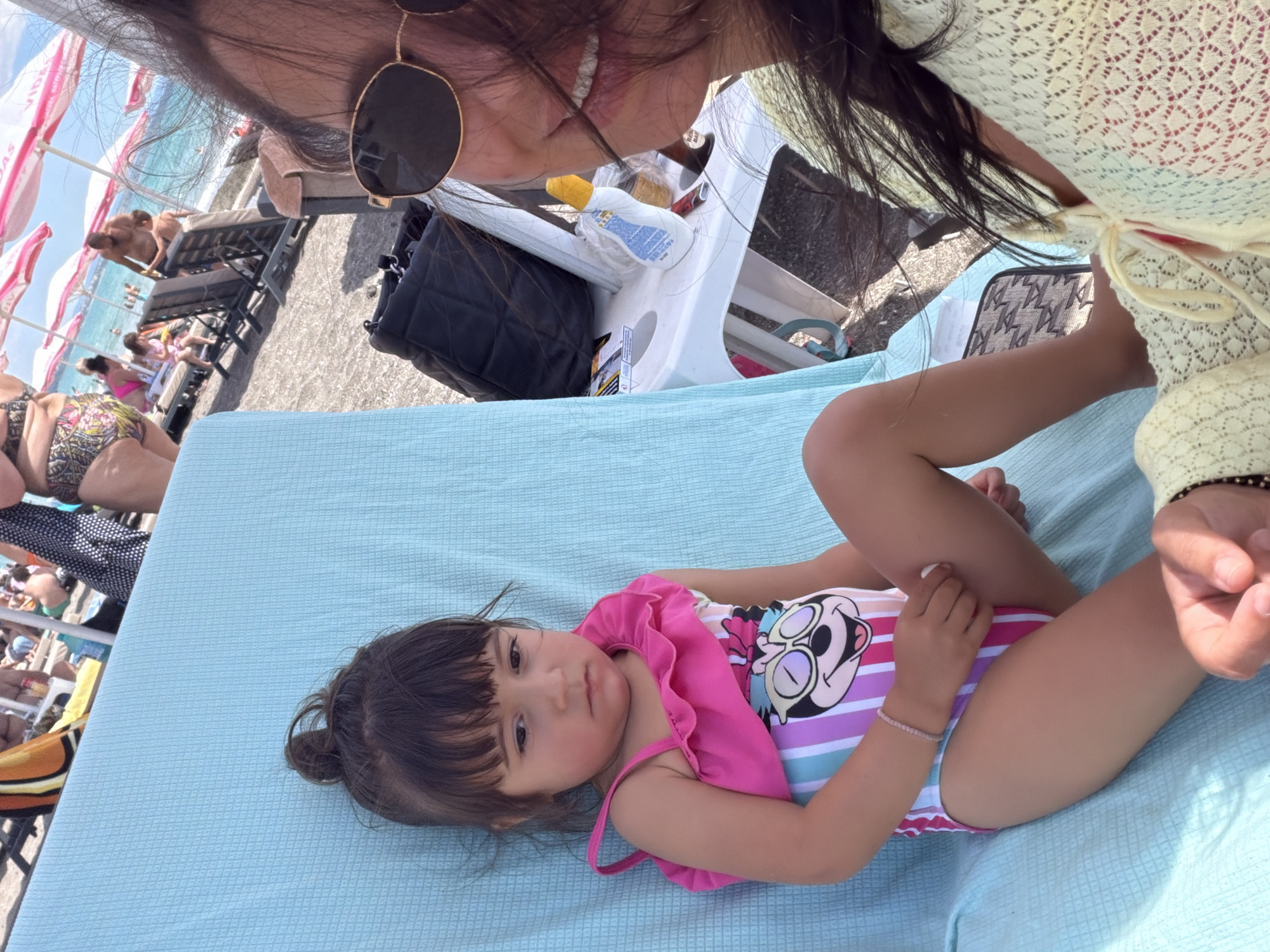 The image depicts a close-up of a young girl and a woman at a beach. The girl, with her hair in a bun and wearing a pink and striped swimsuit with a Minnie Mouse design, is seated on a light blue beach chair. Her expression is somber. The woman, partially visible in the foreground, wears sunglasses and a light yellow crocheted cover-up. She is smiling and her hair is blowing in the wind. In the background, a crowded beach scene unfolds with people relaxing on lounge chairs under umbrellas, and the sea is visible. The location is implied to be Burgas, Bulgaria, a city known for its beaches. The overall setting is a sunny day at the beach, with a focus on the two individuals.