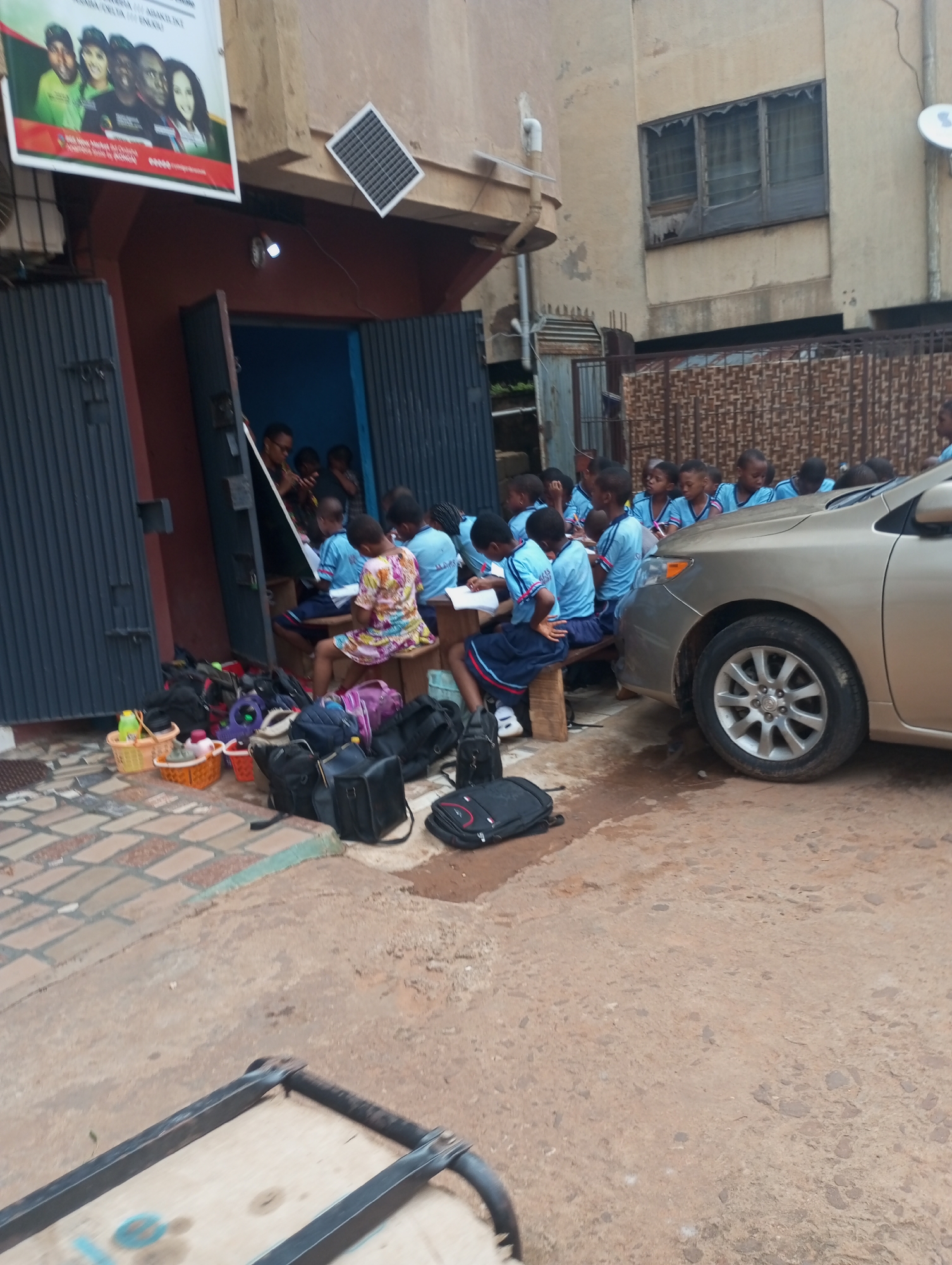 In the city of Onitsha, Nigeria, a group of schoolchildren in blue uniforms are engaged in outdoor learning. They are seated on wooden benches outside a building with a red-painted facade and open metal doors. Some of the children are seen with books and writing materials. A gold-colored car is parked nearby, partially obstructing the view of the children. Scattered on the ground are various backpacks, shopping baskets, and other personal belongings. A poster featuring a group of people is displayed on the wall above the entrance. The ground is paved with interlocking stones in the foreground and transitions to a rougher, dirt-like surface. The overall scene depicts an educational setting taking place outdoors, possibly due to space constraints or a specific teaching method.