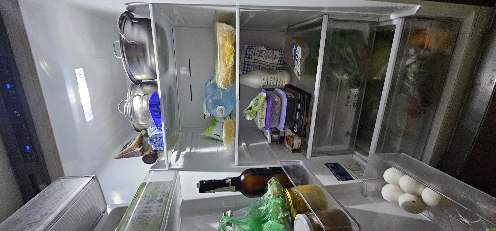 The image shows the interior of a refrigerator, viewed from above. The refrigerator is organized into several compartments. On the left, there are two large metal pots stacked on top of each other. Next to them, there are some food items wrapped in plastic, including what appears to be cheese. Further to the right, there is a large blue water jug, and next to it, more food items in plastic packaging and containers. One container has purple lid and is filled with green leafy vegetables. In the middle section, there is a brown bottle, possibly containing a beverage, lying horizontally. Next to it, a green plastic bag is visible, along with other food items in clear containers. On the far right, there is a clear plastic drawer containing several white eggs. The refrigerator is illuminated by internal lights, which are on. There is no visible indication of the time of day, weather, or emotions. The location is stated as Vinnytsia, Ukraine. Some visible text on the containers and packaging is not legible due to the image quality and resolution.