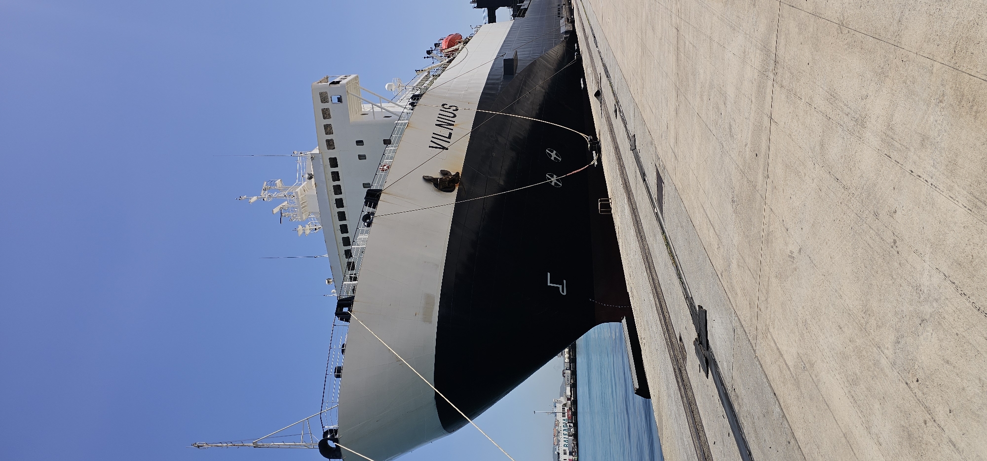 A large, multi-deck ferry or cargo ship, identified by the name "VILNIUS" in prominent white letters on its side, is depicted moored alongside a concrete pier. The ship's hull is painted black below and white above, with a complex white superstructure rising several levels towards the sky. Various antennas, radar arrays, and communication equipment are visible on the ship's mast and upper deck, indicating a functional vessel. A large, dark anchor is secured on the bow, and multiple thick mooring lines stretch from the ship's side to bollards on the pier, holding it in place.
The perspective is low-angle, looking up at the ship, which is angled across the frame. The concrete pier fills the right side of the image, showing textural details, some faint marks, and what appear to be embedded rail tracks. The water, a clear turquoise, is visible in the lower right corner, with part of another, smaller ship, possibly a ferry with "BALEARIA" visible on its side, in the distance. A white numeral "4" is painted on the ship's black bow section near the waterline.
The scene takes place during bright daylight, under a clear, cloudless blue sky, suggesting sunny and fair weather. The overall setting is a bustling commercial port, specifically Algeciras, Spain, given the provided context. No people are explicitly visible, and no particular activity beyond the ship being docked is evident.