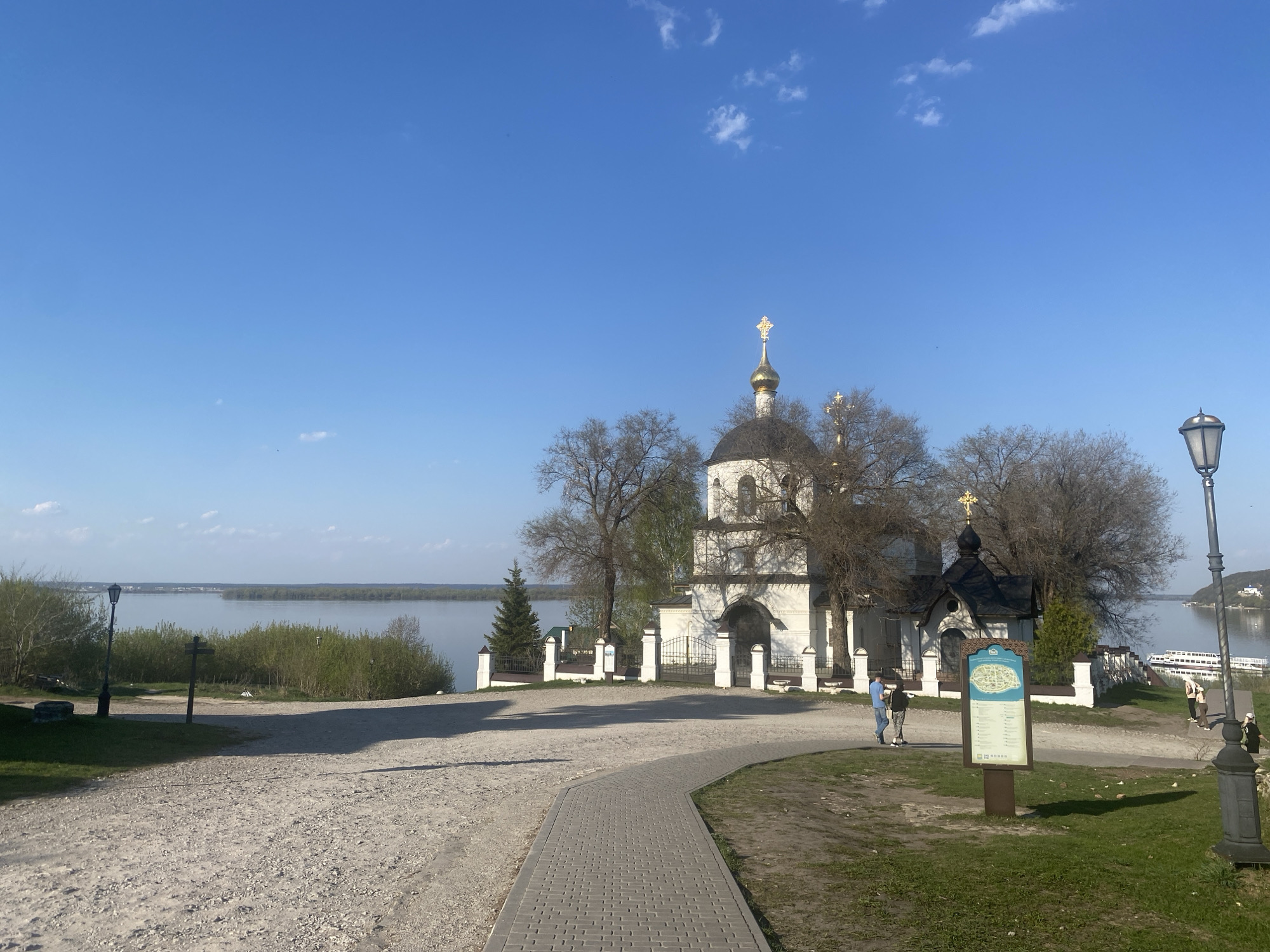 The image shows a view of the Church of St. Sergius of Radonezh in the city of Sviyazhsk, Russia. The church is a white, two-story structure with a black dome topped with a golden cross. It is surrounded by trees with bare branches, suggesting it is either early spring or late autumn.
A paved path leads towards the church, flanked by a dirt and gravel area on the left and a grassy area with a lamp post on the right. A signpost stands near the path. Two people are standing near the signpost, appearing to be looking at it.
In the background is a body of water, likely a river or lake. Vegetation can be seen along the shoreline. The sky is clear blue with a few scattered clouds.
