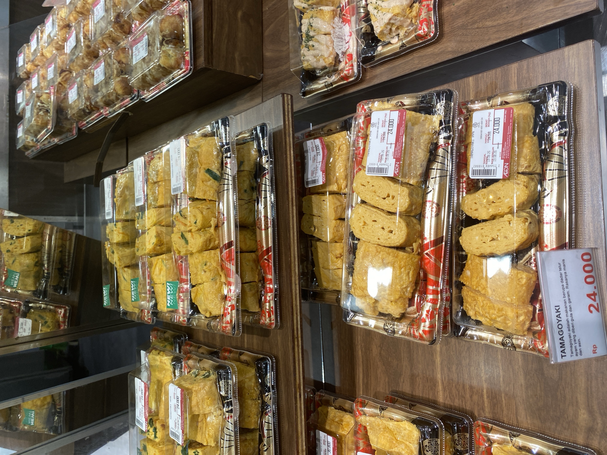 The image shows a close-up, eye-level, interior shot of food items displayed on shelves in a retail setting, likely a supermarket or convenience store, in East Jakarta, Indonesia. The primary subjects are pre-packaged tamagoyaki, a Japanese rolled omelet.
The tamagoyaki is presented in clear plastic containers stacked on wooden shelves. The items are golden yellow, with some variations in color indicating different cooking or ingredient variations, such as visible green herbs in some portions. Each container has a label with product details, including "TAMAGOYAKI", "PPN", "TANGGAL KEMAS 26", "HARGA", "BERAT", and a price in Rupiah, with "24.000" being prominently visible on multiple labels. Some containers also feature "NEW!" stickers. The packaging exhibits red and gold decorative patterns.
In the background, other food items are visible, including more tamagoyaki in similar packaging and other indistinct food products. The lighting is bright and consistent, illuminating the products for display. The environment appears clean and organized, typical of a food retail display area. No people are visible in the frame.