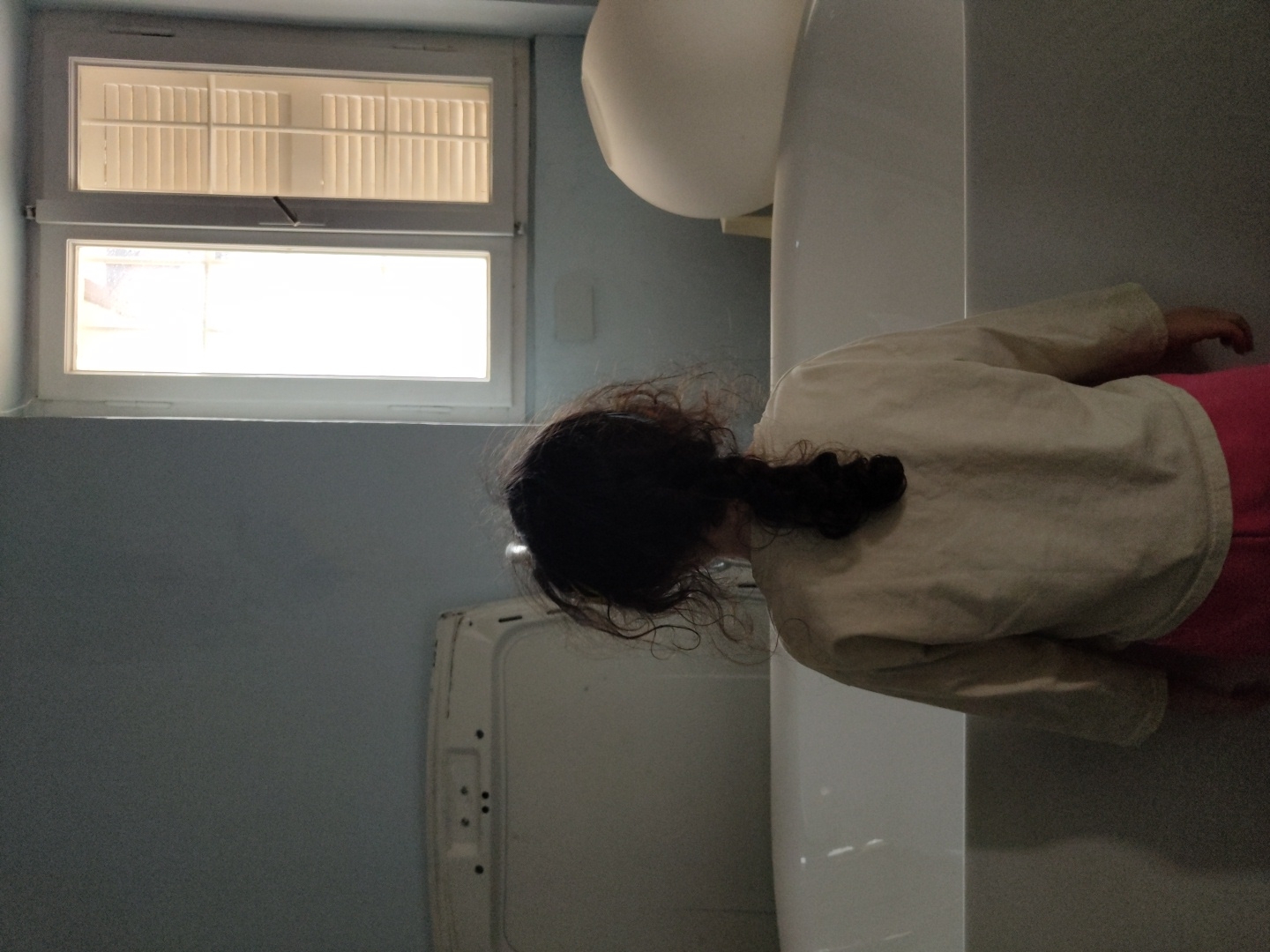 A sideways, medium shot shows a bathroom interior. A young person wearing a beige top and pink bottoms is positioned at the right side of the image next to a white toilet and a long, narrow white sink. The person's back is to the camera, and they have dark curly hair tied back in a small ponytail. To the left, the wall is painted a light blue color. Above is a rectangular window with white frame. The top section of the window has light-colored horizontal blinds, and the bottom section has clear glass, letting in bright light. Below the window to the left is a white object resembling a clothes washer. The location is Unknown, Unknown.